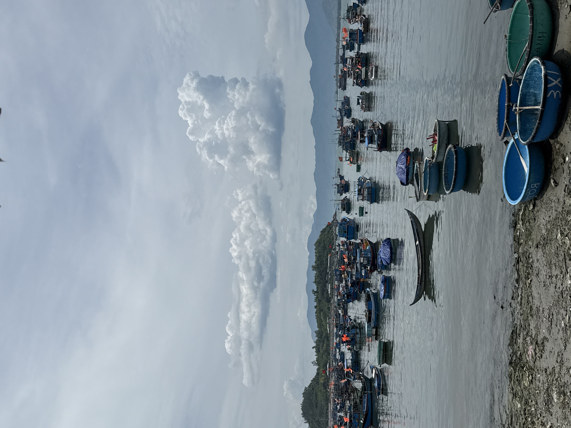 The image depicts a large fleet of fishing boats anchored in the water near a shore. The boats vary in size and color, with many being blue and some featuring orange flags. In the foreground, several round, basket-like boats are pulled up onto the muddy shore. The background shows a lush, green coastline with hills and mountains under a cloudy sky with a prominent, large cumulus cloud formation. The scene appears to be a harbor or bay, likely in Vĩnh Lương, Vietnam, given the prevalence of fishing vessels and the tropical landscape. There are no people visible in the frame. The water is calm, reflecting the sky and boats. The shore is covered with debris, suggesting a natural or semi-developed coastline.