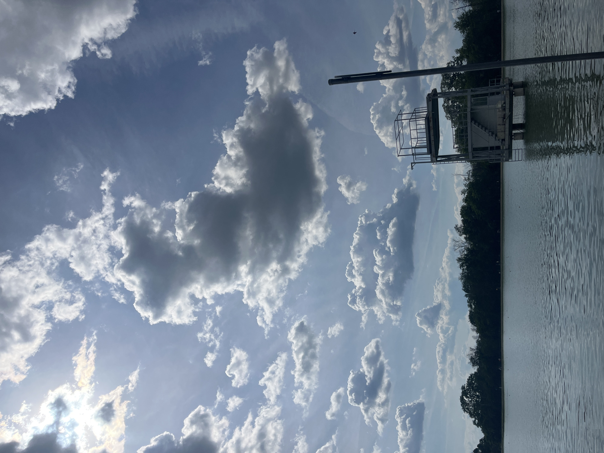 The image shows a lakeside landscape in Plovdiv, Bulgaria. To the right is a metal diving platform structure rising above the water. Adjacent to the water's edge is a line of trees, dark against the bright sky. The sky dominates the view, filled with white cumulus clouds scattered across a blue background. A thin pole or beam is visible extending upward from the diving platform. The water appears relatively calm, reflecting the sky.