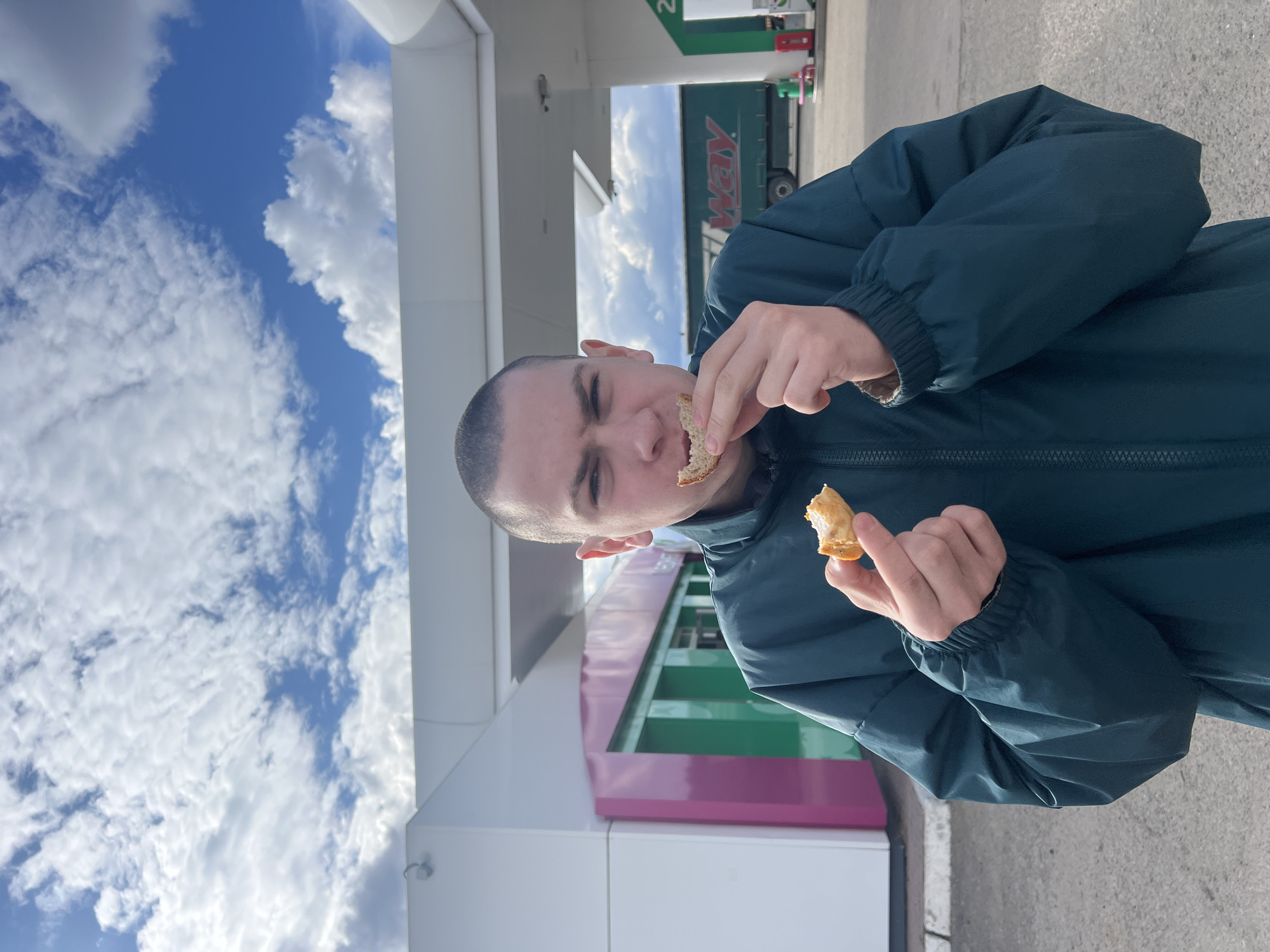 The image captures a man eating outdoors at what appears to be a gas station or service area in Alnashi, Russia, during bright daylight. The sky is a vivid blue with numerous white, fluffy clouds.
The central subject is a man with a shaved head, wearing a dark teal or deep green hooded jacket. He is actively eating, holding a piece of light-colored bread or pastry in each hand; one piece is partially bitten and held close to his mouth. His expression is focused as he consumes the food.
In the background, to the left of the man, are modern-looking building structures with white, pink/purple, and green sections. To his right and further back, under a white canopy, a large green truck with white lettering "AY" (likely part of a word like "WAY") is visible. A green sign displaying the number "2" can also be seen, possibly indicating a pump or service bay. The setting suggests a roadside stop for fuel and refreshments.