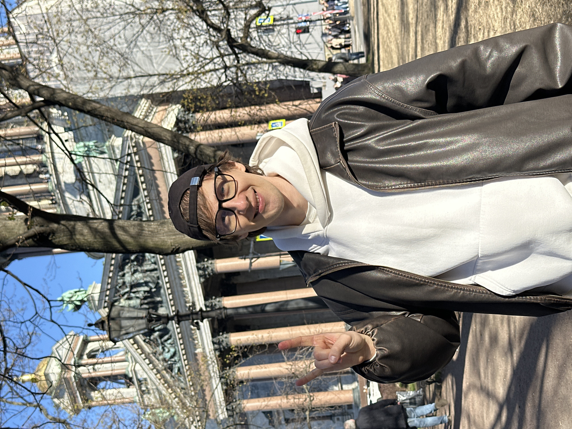 A young man is smiling at the camera, making a "rock on" hand gesture, and standing on the sidewalk in front of Saint Isaac's Cathedral in Saint Petersburg, Russia. He is wearing glasses, a dark-colored baseball cap, a white hoodie, and a dark-brown leather jacket. Behind him, the massive Saint Isaac's Cathedral is partially visible, with its iconic columns, sculptures, and dome. Bare tree branches are visible against a blue sky. People are walking on the sidewalk in the background.