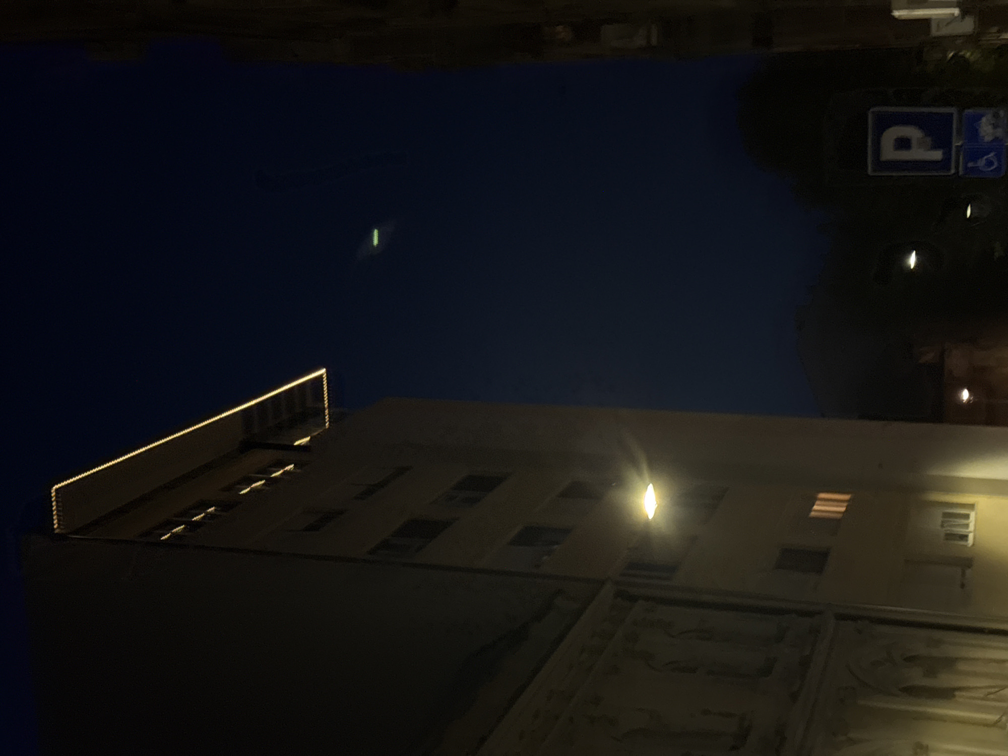 The image shows a building at night in Belgrade, Serbia. The building is lit by a streetlight and appears to have lights along the roofline. To the right of the building, there's a parking sign. The sky is dark, with a faint green light visible in the upper left.