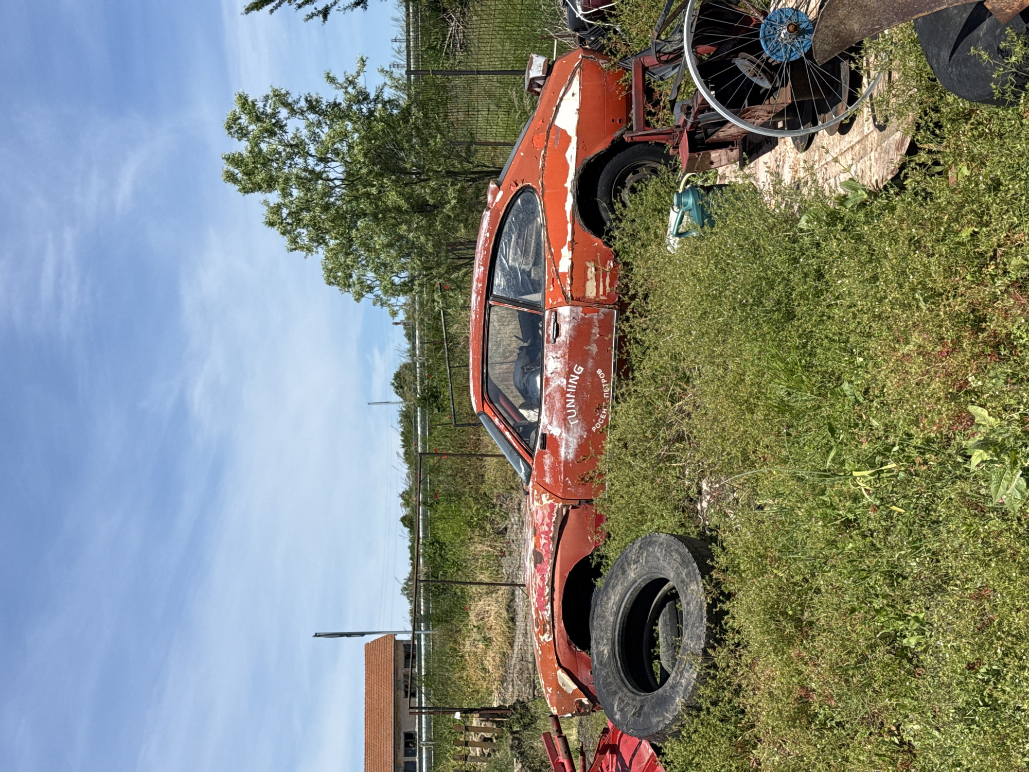 The image shows a rusty, partially disassembled red car in an overgrown field. The car appears to be in a state of disrepair with missing parts and faded paint. The word "TUNNING" is visible on the car's body. A tire rests near the front of the car. The background features a fence, a tree, and a blue sky. The location is Omurtag, Bulgaria.