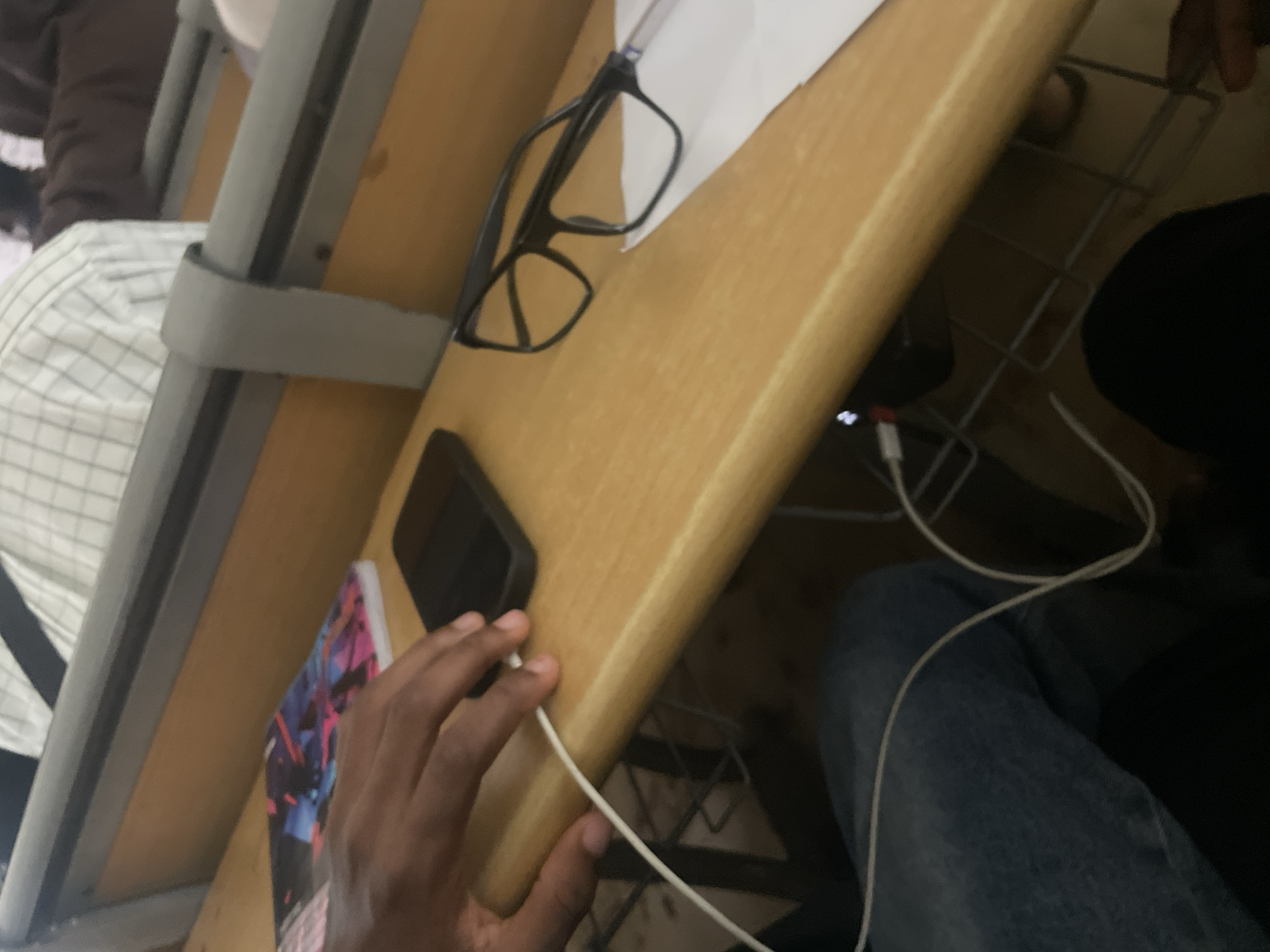 A person's hand, with dark skin, rests on a wooden desk in a classroom setting in Ihiagwa, Nigeria. The hand is positioned near a black smartphone and a notebook with a colorful, abstract design. A white charging cable extends from the phone, looped around the person's wrist and disappearing out of frame. On the desk, a pair of black-rimmed glasses sits on a piece of white paper. In the background, a student wearing a plaid shirt is visible. The desk is part of a larger piece of classroom furniture, with metal supports and gray straps visible. The overall environment appears to be a typical academic setting, likely a lecture hall or study area.