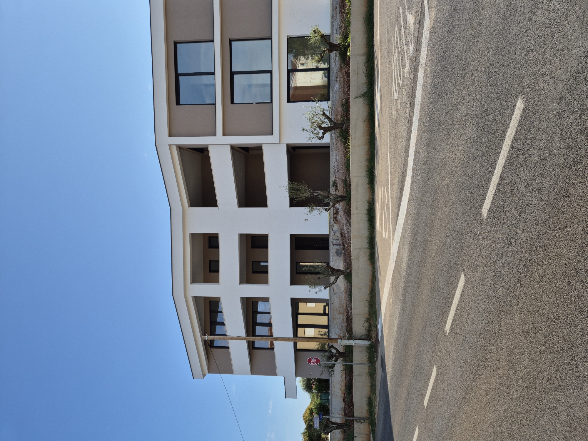 The image shows a modern building with a beige and white facade, featuring multiple balconies and large windows. The building is located in Zambratija, Croatia, as indicated by the street sign on the left. In front of the building, there is a road with white lane markings and a stop sign. Several small olive trees are planted in front of the building, between it and the road. The sky is clear and blue, and the overall setting appears to be a sunny day. There are no people visible in the image.