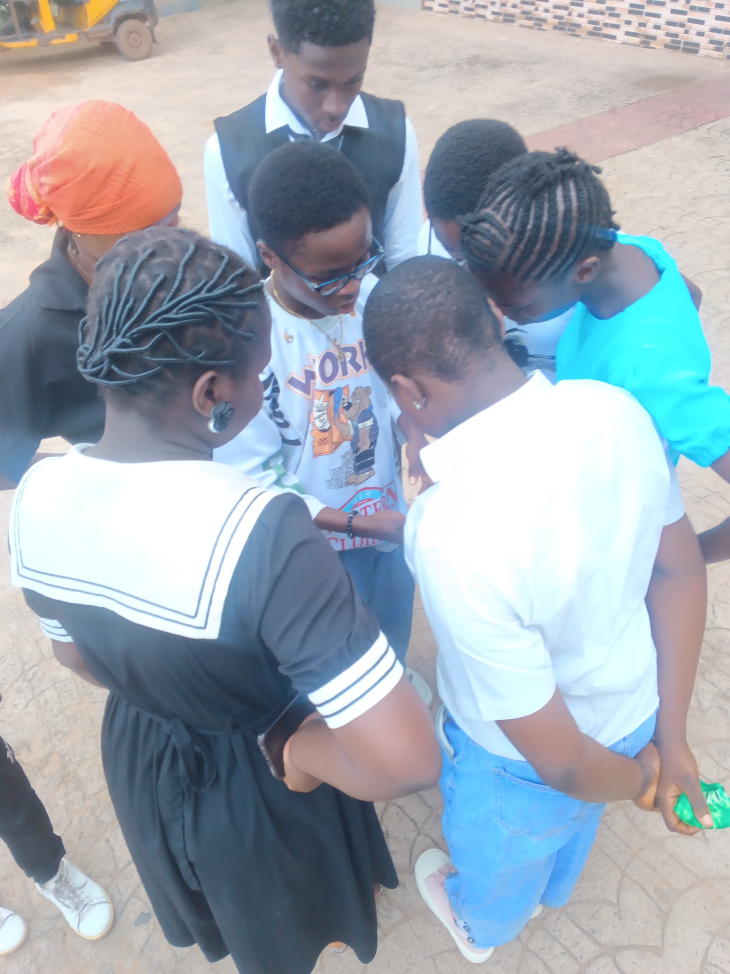 The image depicts a group of seven individuals gathered in a circle outdoors on a paved surface, possibly in Nkpor, Nigeria. The people are of varying ages and appear to be looking down at something in the center of the circle, suggesting a shared activity or discussion.
The individuals are dressed in a mix of casual and somewhat formal attire. One person on the far left wears an orange headscarf and a black top. Another person in the center foreground wears a black and white dress with a sailor-style collar. To their right, a person in a white t-shirt and blue jeans is holding a green object. Behind them, another individual is wearing a blue shirt. In the background, a young man is visible wearing a white shirt and a black vest. The other individuals' faces are partially obscured or turned away, making detailed descriptions difficult.
The ground is a patterned pavement, and in the background, a blurry yellow and black vehicle, possibly a tricycle or a small van, is visible. The overall lighting suggests it is daytime. The scene appears to be public, possibly a street, plaza, or school compound.