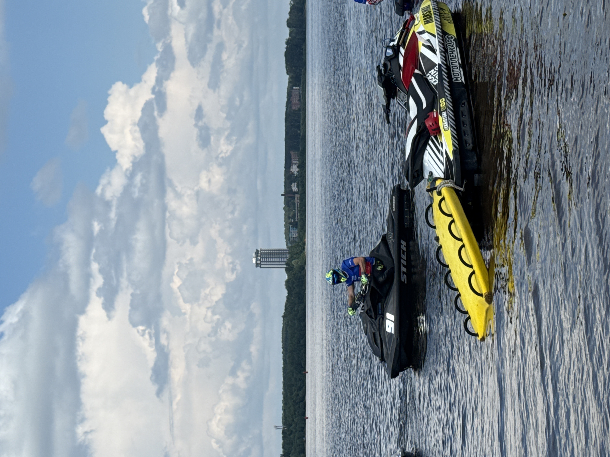 In the city of Perm, Russia, on a body of water, a jet ski race is in progress. The image captures several jet skis on the dark blue water, under a partly cloudy sky. In the foreground, a black jet ski with the number "91" is being ridden by a person wearing a blue shirt and a yellow and blue helmet. Next to it, a second jet ski, predominantly black, white, and red, with various decals including "MARSHAL" and Cyrillic lettering, is stationary. A yellow flotation device with black handles is attached to this jet ski. In the background, a treeline borders the water, with buildings and a tall, modern skyscraper visible on the horizon, indicative of a city. The water is rippled, suggesting movement or a light breeze.