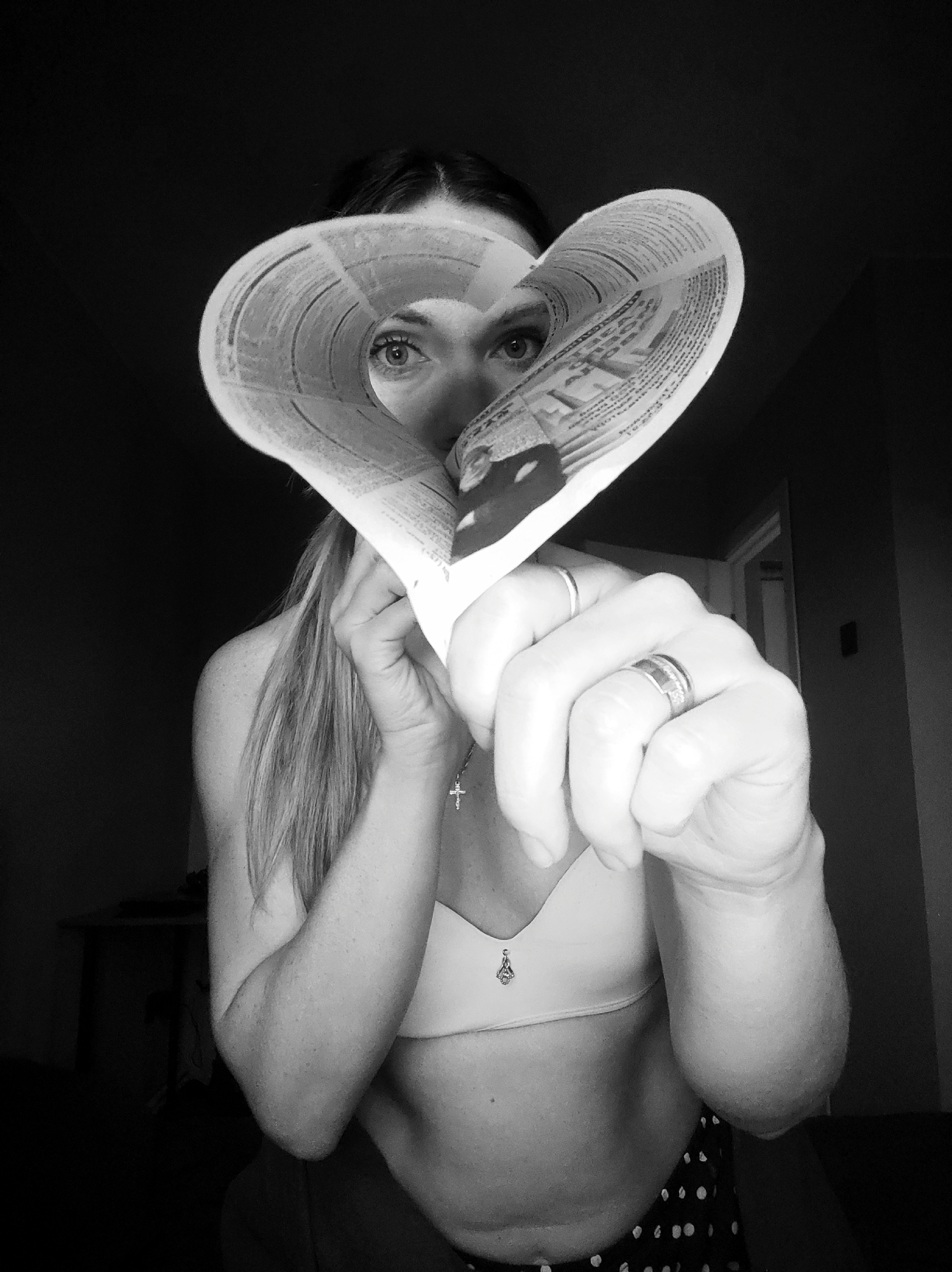 This black and white portrait features a woman indoors, looking directly at the viewer through a heart-shaped paper cutout. Her eyes are prominent and engaging, framed by the edges of the paper. She holds the paper heart with both hands, her left hand extended towards the camera, prominently displaying two rings: one on her ring finger and another on her pinky. A small cross pendant necklace is visible around her neck.
The woman has long, dark hair, partially draped over her left shoulder. She is wearing a light-colored bra and what appears to be polka-dotted shorts or pants. The paper heart is made from printed material, likely a newspaper or magazine, with visible but illegible text and small images on its surface.
The scene is dimly lit, creating a low-key aesthetic that highlights the woman's face, hands, and the paper heart against a dark, out-of-focus background. Hints of a doorframe and a table or desk are discernible in the shadows. The direct gaze through the heart shape conveys a sense of intimacy or connection. This scene takes place in Votkinsk, Russia.