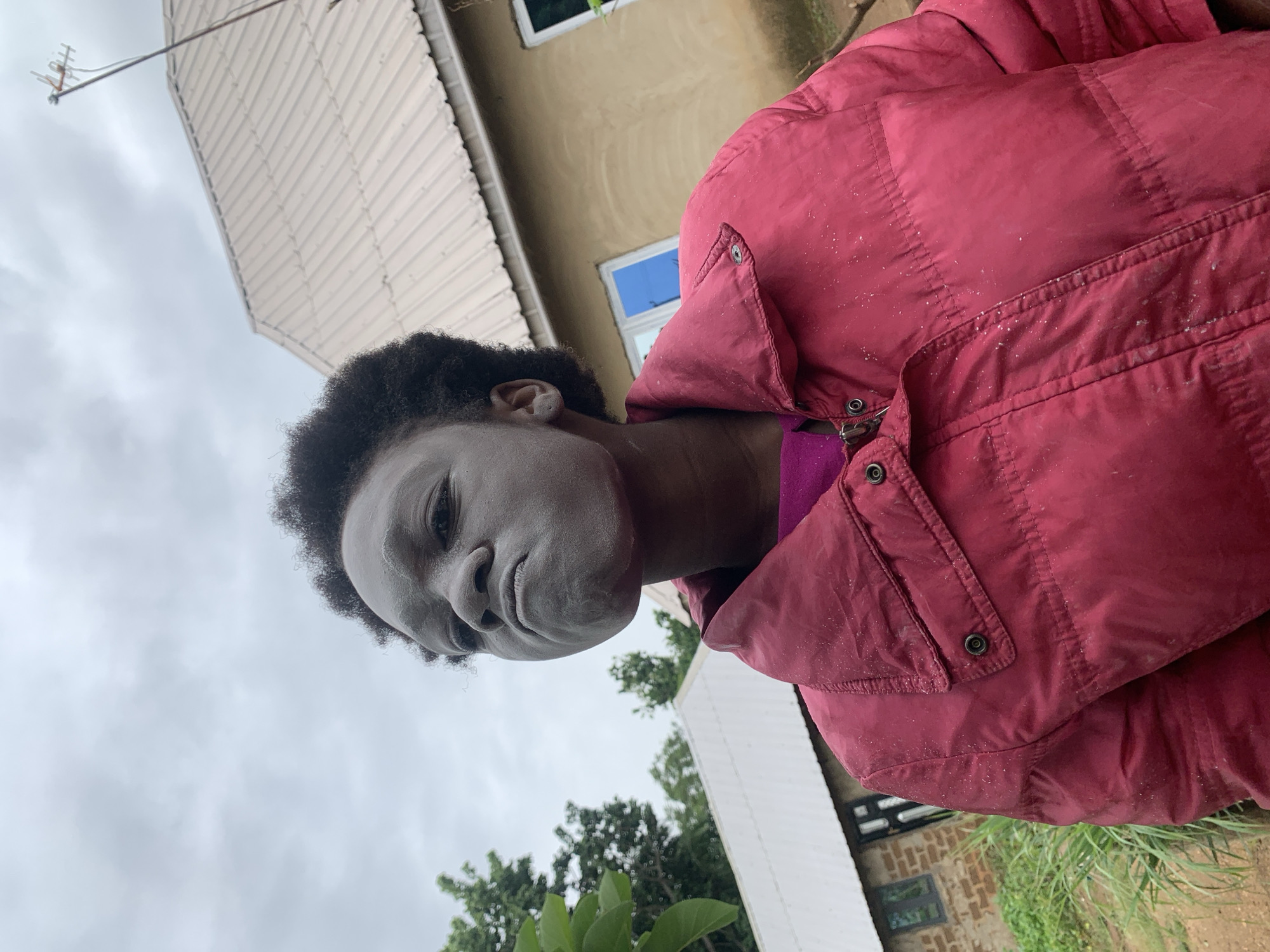 A medium shot captures a person with dark curly hair and a face covered in white powder. The person is wearing a maroon-colored puffy jacket over a purple shirt. The background includes parts of buildings: one with beige stucco walls and blue-framed windows, and another with a white corrugated metal roof. Green trees and plants are visible near the bottom of the image. The setting appears to be outdoors in Agban, Nigeria.