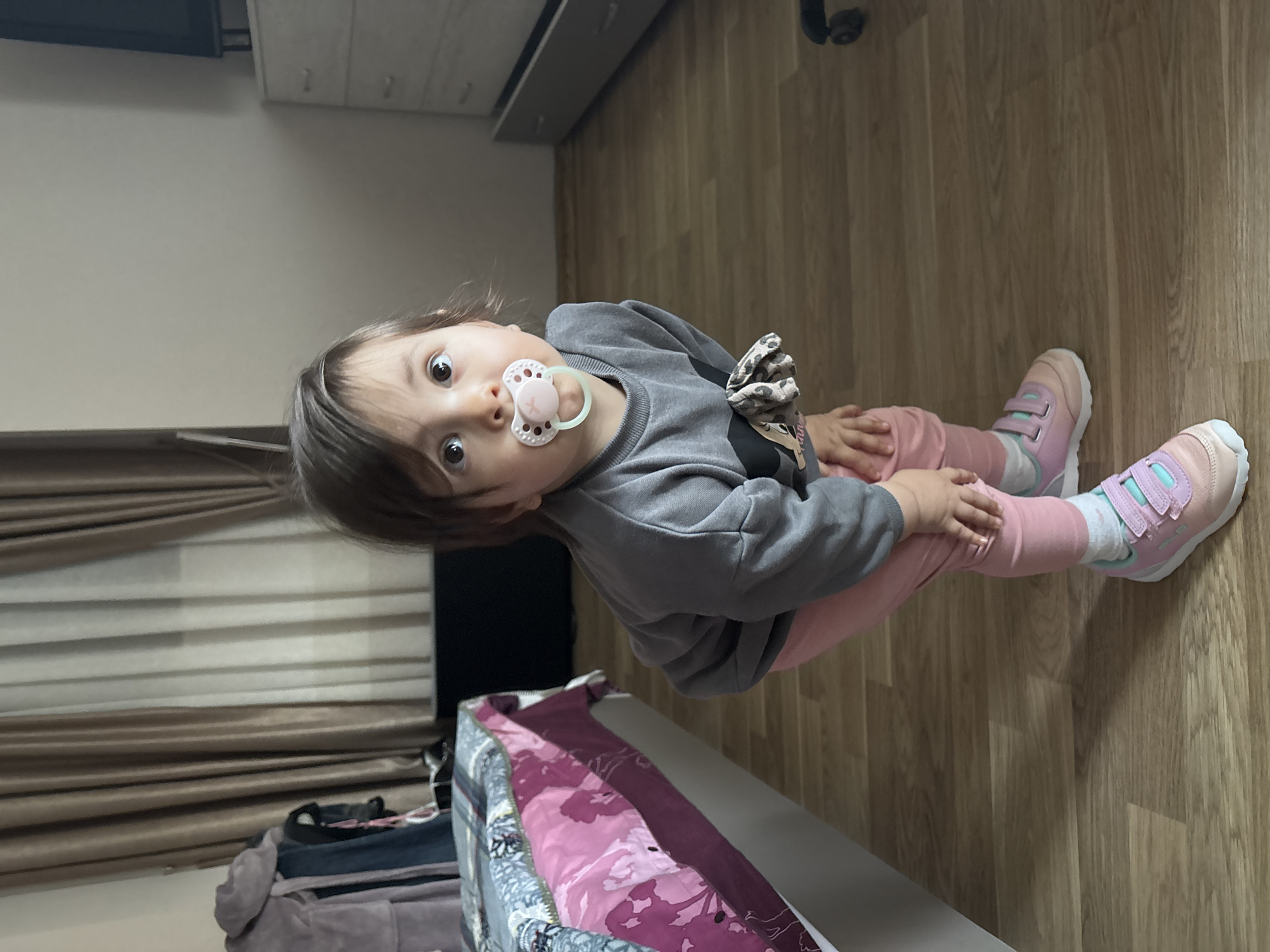 A young child with a pacifier sits on a wood floor in a room in Plovdiv, Bulgaria. The child wears a gray sweatshirt with a decorative bow, pink pants, pink and lavender shoes, and white socks. A partially visible object with purple and white fabric rests nearby, possibly a bed or changing table. In the background are a dresser and a window with closed blinds.
