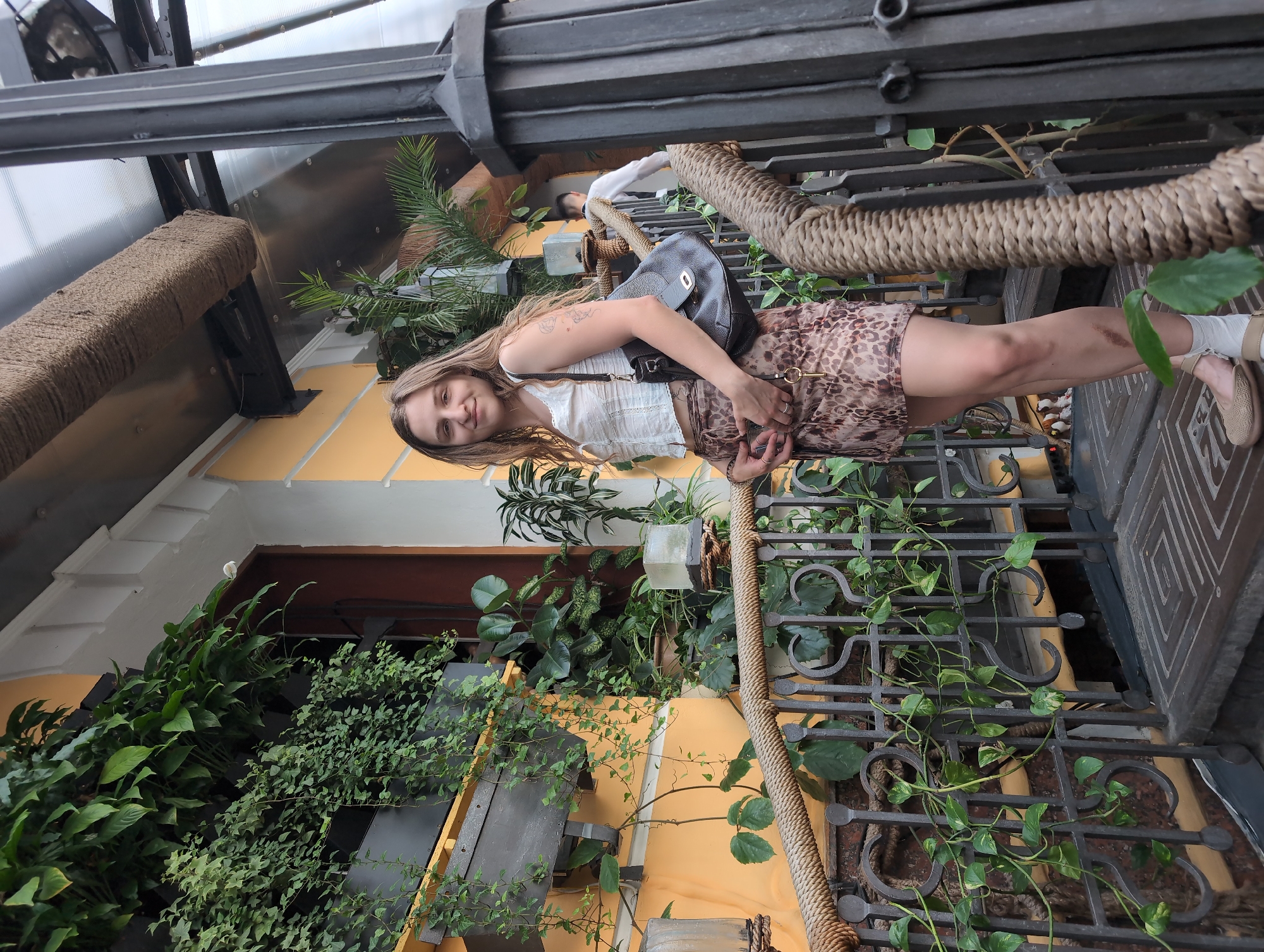 A young woman with long, wavy brown hair stands on a metal staircase adorned with a thick rope railing and overflowing with green plants. She is wearing a white, cropped, sleeveless blouse with a lace trim and a short, leopard-print skirt. A dark brown leather handbag hangs from her right shoulder. Her left hand rests on her stomach, holding a smartphone, while her right hand is down by her side. She is wearing beige flat shoes with white socks. The staircase is situated in front of a building with a pale yellow facade, surrounded by various potted and hanging plants, creating a lush, green environment. In the background, another person in a white shirt and dark vest is walking up the stairs. The scene appears to be an outdoor seating area or an entrance to a cafe or restaurant in Kyiv, Ukraine, possibly on Andriivskyi Descent, known for its artistic atmosphere and numerous cafes.