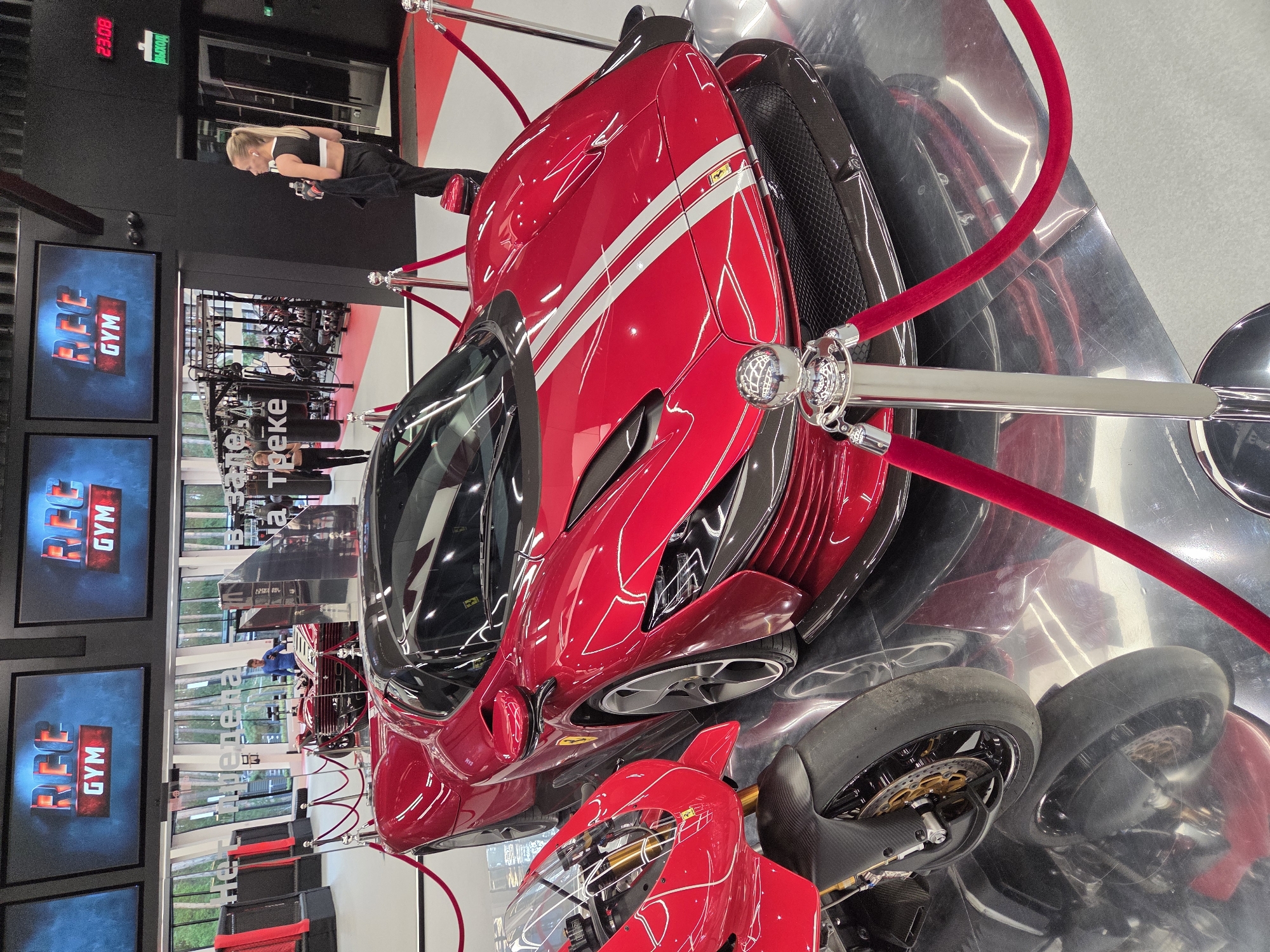 A red Ferrari sports car and a red motorcycle are on display in an indoor exhibition space. The car is in the foreground, angled towards the right. It is a low-profile vehicle with a sleek design, featuring white racing stripes on its hood. The motorcycle is to the left of the car, partially obscured. Both vehicles are positioned on a highly reflective floor. Red velvet ropes on stanchions create a barrier around the display. In the background, there are large screens displaying the "REC GYM" logo. A woman in athletic wear is standing to the right of the car, holding a water bottle and a towel. Behind the vehicles, gym equipment is visible through large windows. The overall environment suggests a modern fitness facility or a promotional event. This scene is located within the REC GYM in Yekaterinburg, Russia.