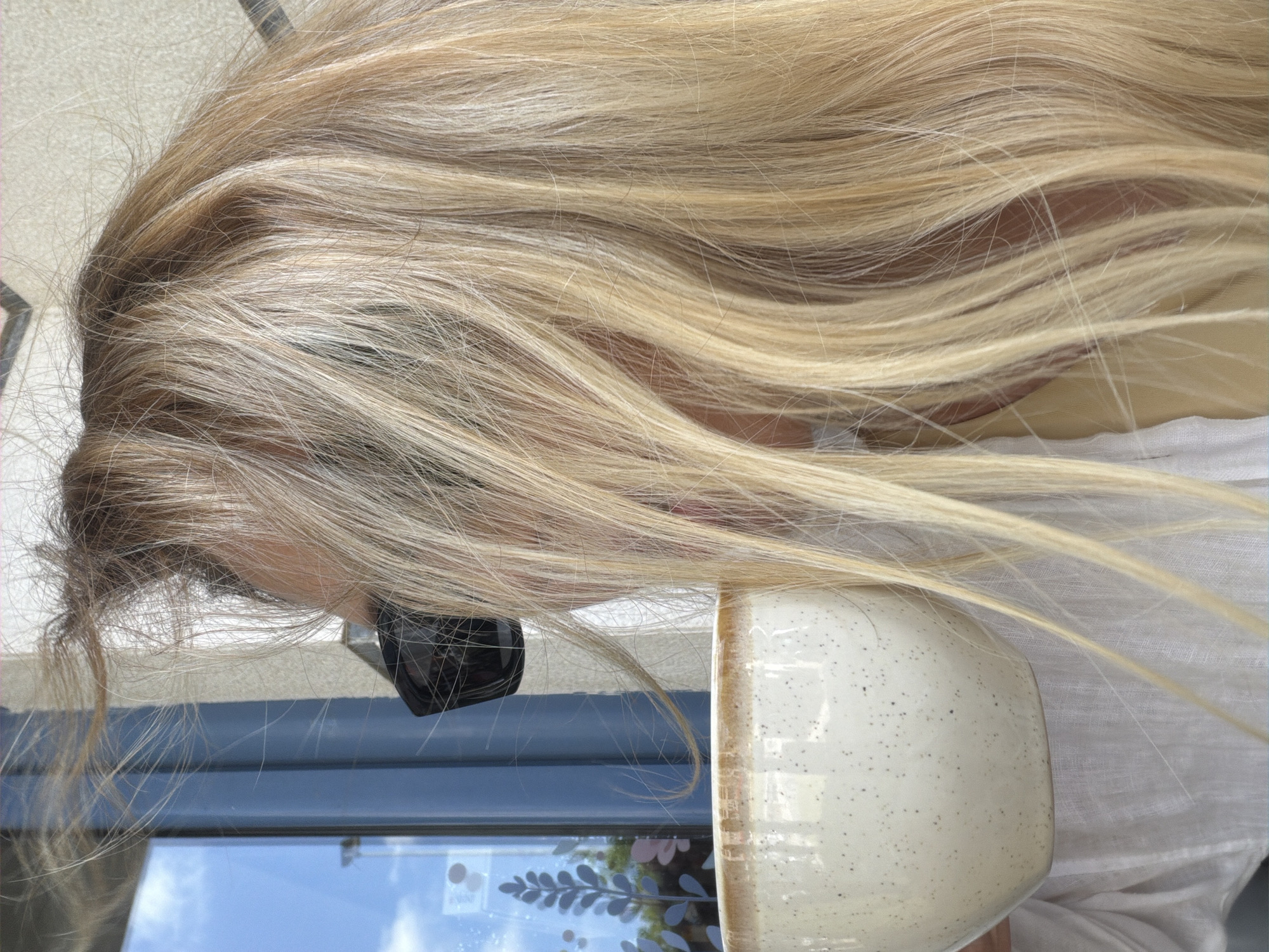 The image shows a close-up of a woman's head and shoulders as she holds a speckled ceramic mug with a light brown rim. She is wearing sunglasses and a white linen top. Her long, blonde hair is partially obscuring her face and is blowing to the left. In the background, a blue window with a decorative plant motif is visible. The scene appears to be outdoors, possibly at a cafe in Budva, Montenegro.