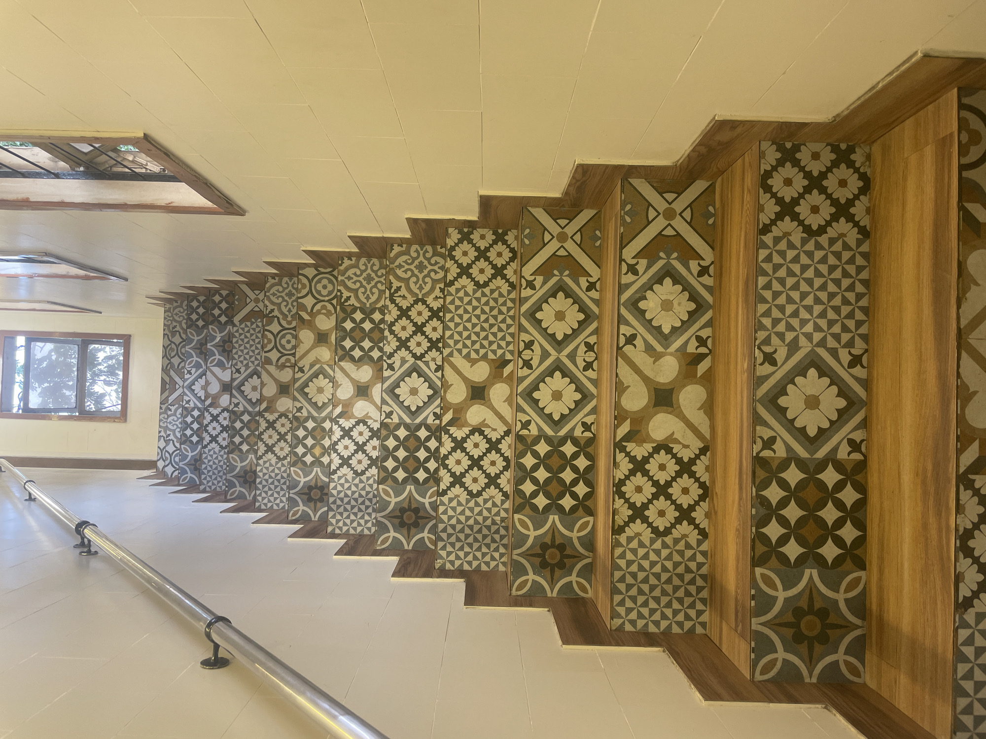 The image depicts a unique stairwell. The stairs are painted white, with a series of intricately patterned tiles installed vertically along the wall. Each tile row is framed by wooden trim. A silver handrail runs along the left side of the stairs. A window is visible at the far end of the stairwell, providing natural light. The ceiling is white and flat. Based on the architectural style and design elements, it is highly plausible that this is an interior shot taken in Mumbai, India.