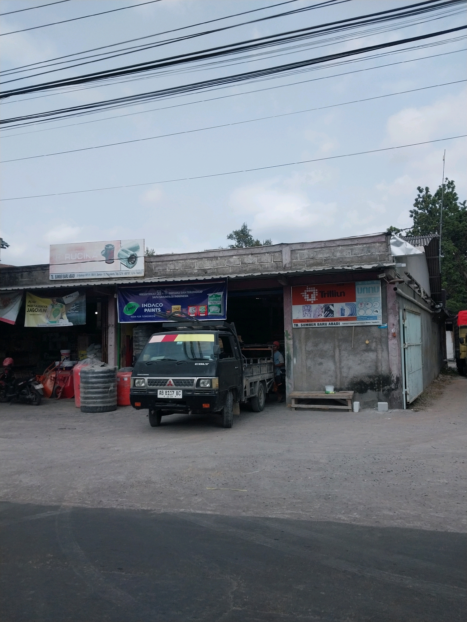 The image captures a storefront on a cloudy day in Purwomartani, Indonesia. The building appears to be a retail establishment selling building materials and paint, indicated by banners and signage for brands like "RUCIKA," "INDACO PAINTS," and "Trillium." A black Mitsubishi Colt pickup truck, license plate "AB 8117 BC," is parked in front of the store, partially obstructing the entrance. To the left of the truck, a red motorcycle is parked. Several large cylindrical objects, possibly water tanks or storage containers, are stacked outside the store. Inside the open storefront, shelves with products and more storage units are visible. A person, wearing a red shirt, is standing near the entrance. The building's facade is constructed with rough concrete blocks and has a corrugated metal roof. Overhead power lines crisscross the sky, and a few trees are visible in the background. The foreground shows a paved road and sidewalk.