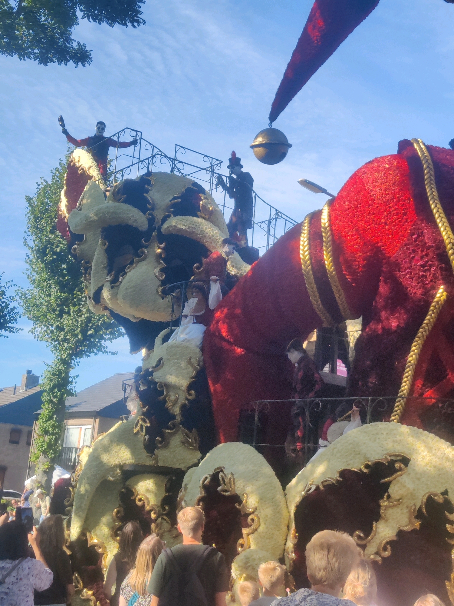 This is a medium shot, taken from a low angle, of a large, elaborate float decorated with flowers, likely for a parade. The float depicts a giant jester's head, with a prominent white and gold face. Behind the head, a large red section of the float rises, adorned with thick gold braiding. On top of the float, several figures are visible. One figure, dressed in black and red with white face paint, stands on a railing, holding what appears to be a bottle. Another figure, in a dark uniform and hat, stands on a platform. A large golden bell hangs from a pointed red structure extending from the top of the float into the blue sky. The float is positioned on a street, with trees and residential buildings visible in the background, suggesting an urban or suburban setting. The sky is clear and blue. In the foreground, the backs of several people are visible, looking towards the float, indicating they are spectators. The scene is likely from the annual Bloemencorso Zundert, a large flower parade held in the city of Zundert, Netherlands, known for its elaborate floats made entirely of dahlias.