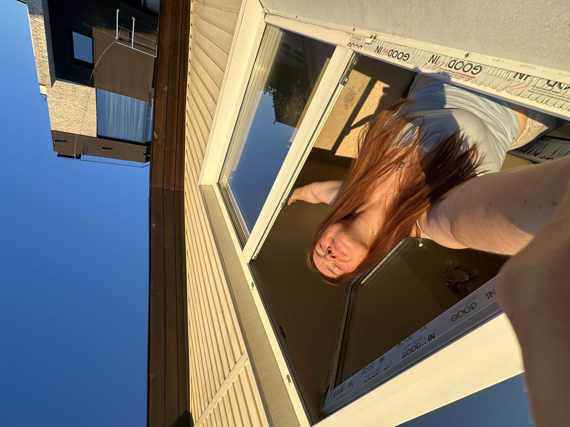 A young woman with long, red hair is leaning out of a window, her face tilted towards the sun. She is wearing a light blue top. The window is open, and the sunlight illuminates her face, creating shadows. The building exterior is a light yellow color with horizontal siding. In the background, a modern building with dark brown and patterned facade elements is visible against a clear blue sky. The photo appears to be taken from a low angle, looking up towards the woman and the buildings. There are no discernible landmarks that place this scene specifically in Nizhny Novgorod, Russia.