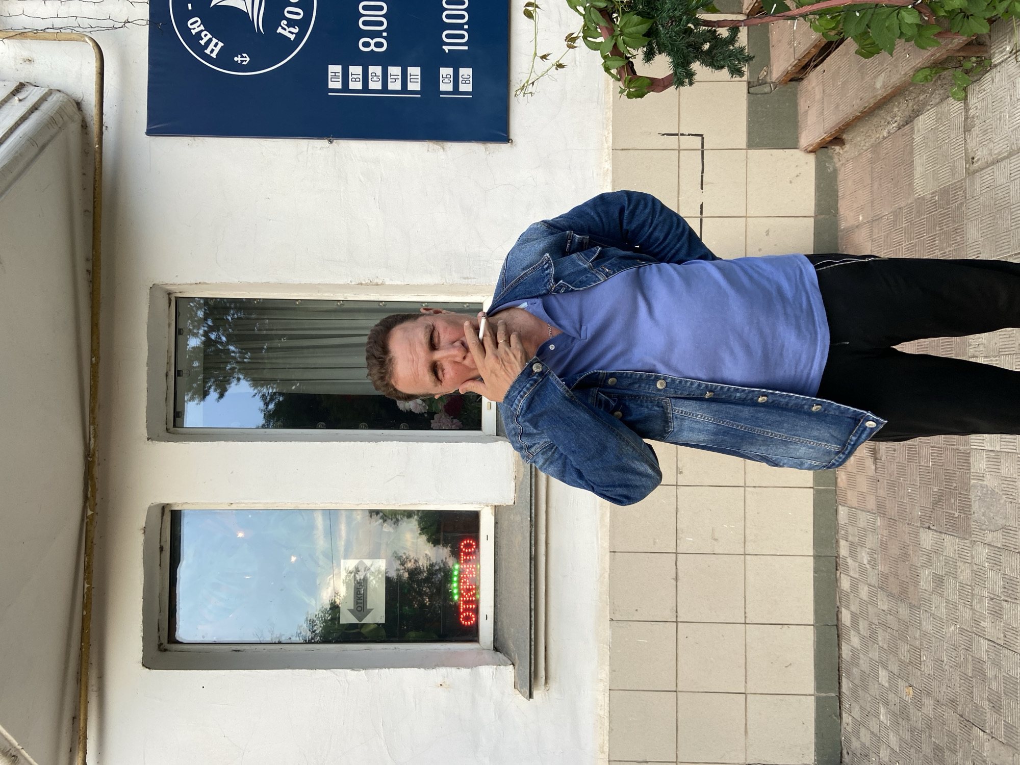 A man in a blue denim jacket and a light blue polo shirt stands outside a building, smoking a cigarette. He is looking at the camera with his left hand holding the cigarette to his mouth and his right hand on his hip. Behind him, there are two windows. The left window has a sign that says "ОТКРЫТО" (OPEN) in red neon letters with green lights above it, and a white arrow pointing to the left. The right window shows a curtain and some plants. To the right of the windows, a dark blue sign with white lettering displays "НЬЮ - КО" and a schedule with days of the week and times. The building has a white, textured facade with light-colored tiled squares below the windows. The ground is paved with square tiles. A small evergreen plant and a wooden bench are visible to the right of the man.