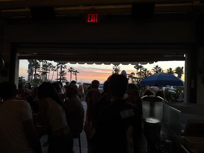 A dim interior frames an outdoor gathering under a sunset sky. Silhouetted figures mingle, palm trees sway against the colorful horizon, and a large blue umbrella provides shade. An "EXIT" sign glows red overhead. The scene captures a relaxed, social atmosphere at dusk, likely at a beachside bar or restaurant. A beverage dispenser stands near a serving area.