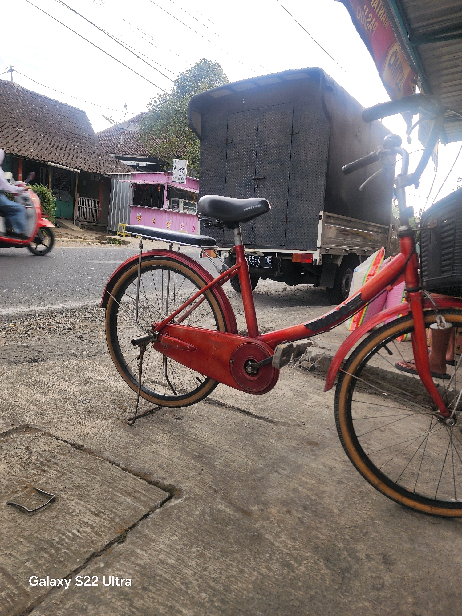 This image captures a street scene in Salaman, Indonesia. In the foreground, a red bicycle with a black seat and basket is parked on a concrete sidewalk. The bicycle has a kickstand deployed, supporting its weight. Behind the bicycle, a large, dark-colored truck with a diamond-plated rear is parked. The truck has a license plate that reads "8594 DE". To the left of the truck, a red scooter is visible, with a person riding it, wearing blue jeans and a white top. The background shows several buildings, including houses with tiled roofs and a pink storefront. The sky is overcast. The ground is paved with concrete and asphalt, with some loose gravel and debris. The overall impression is of a typical street scene in a moderately populated area.