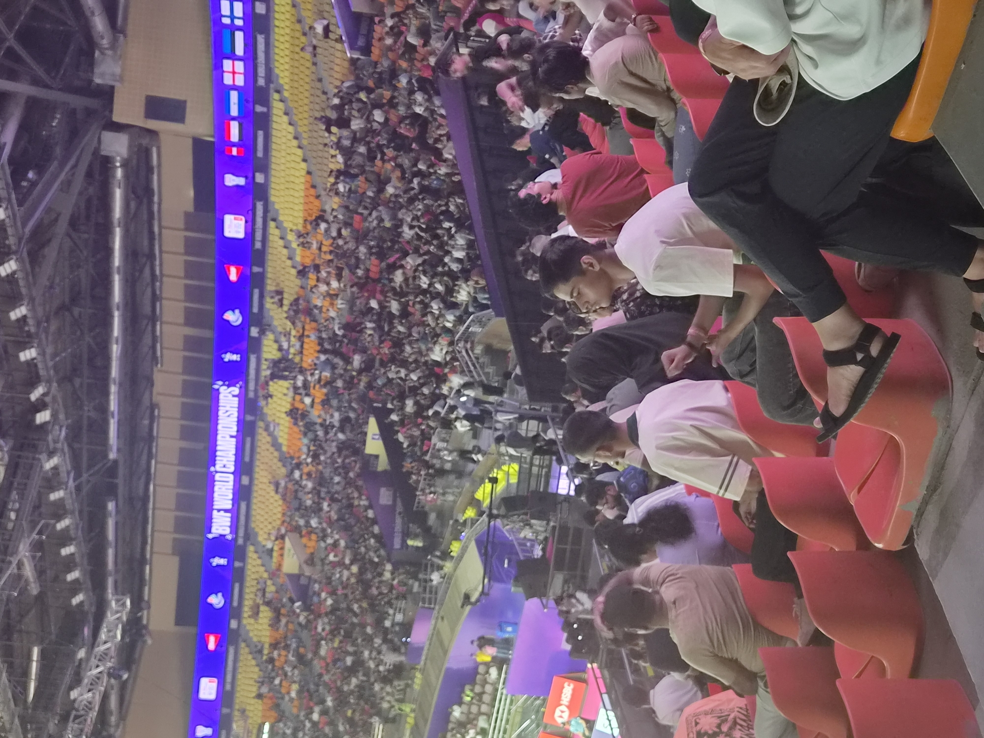 This is a photograph taken in an indoor stadium, likely the Indira Gandhi Indoor Stadium in Delhi, India, during the BWF World Championships. The image captures a section of the audience seated in bright orange stadium seats. The stands are filled with a diverse crowd of people, many of whom are facing towards a stage or court that is not fully visible in the frame. In the upper portion of the image, a large LED screen displays the text "BWF WORLD CHAMPIONSHIPS" along with various country flags and event logos. The stadium architecture, with its tiered seating and exposed lighting and structural elements, is indicative of a large sports venue. The lighting appears to be artificial, typical of an indoor event. The people in the foreground are dressed in casual attire, suggesting they are spectators. Some individuals are looking at their phones, while others are engaged in conversation or watching the event. The overall atmosphere seems to be one of a sporting event with a considerable audience present.