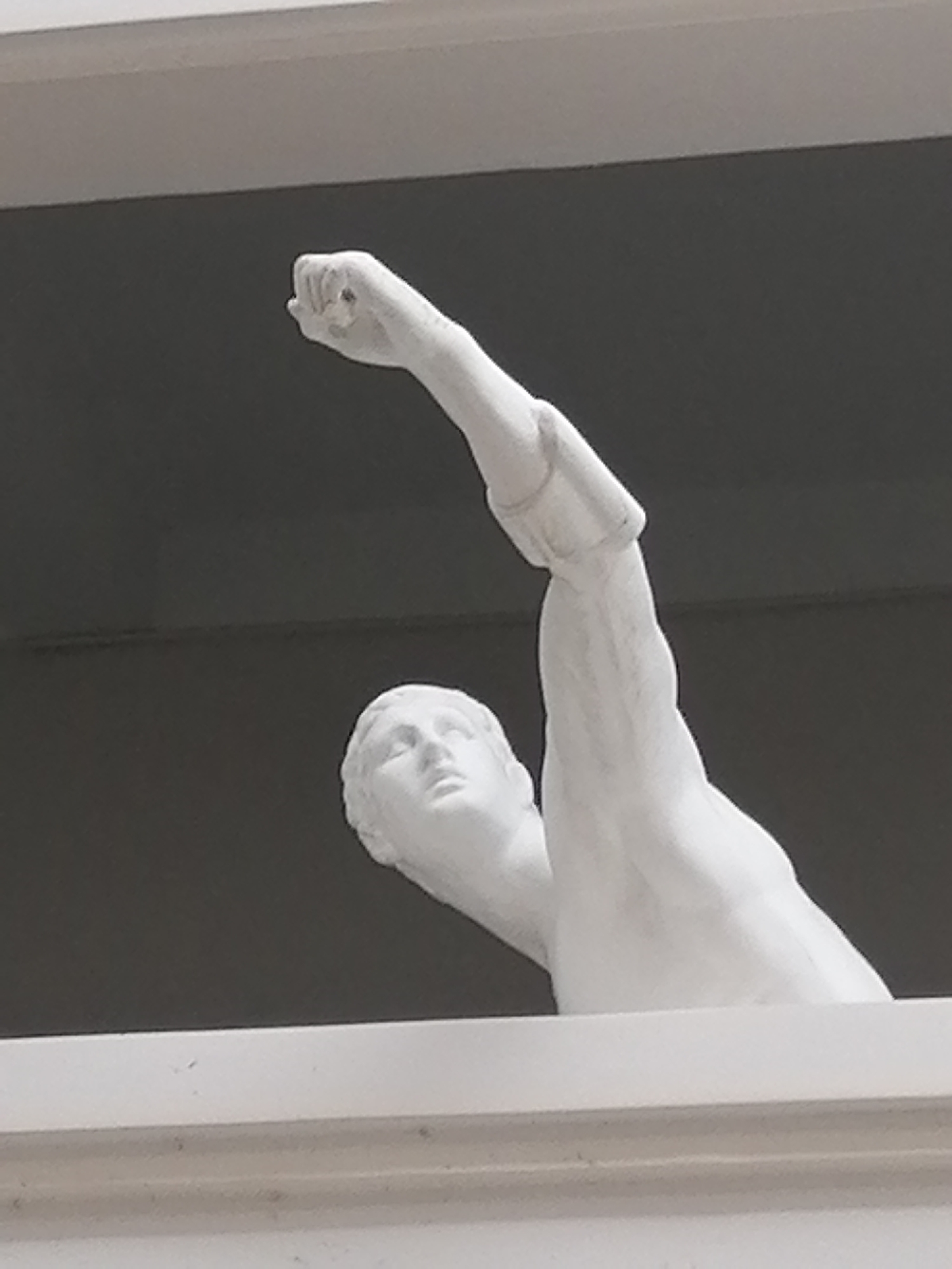 A white stone statue of a man is partially visible, emerging from behind a white ledge. The statue is sculpted in a realistic style, with smooth contours and anatomical detail. The man's head is turned slightly to his left, with his face looking upwards. His right arm is raised and bent at the elbow, with his fist clenched. The statue is positioned outdoors, against a dark gray background, possibly a building or sky. The image is taken from a low angle, looking up at the statue. The white ledge in the foreground is decorative and appears to be part of a building. No specific location in Lâm Viên - Đà Lạt, Vietnam can be determined from the image.