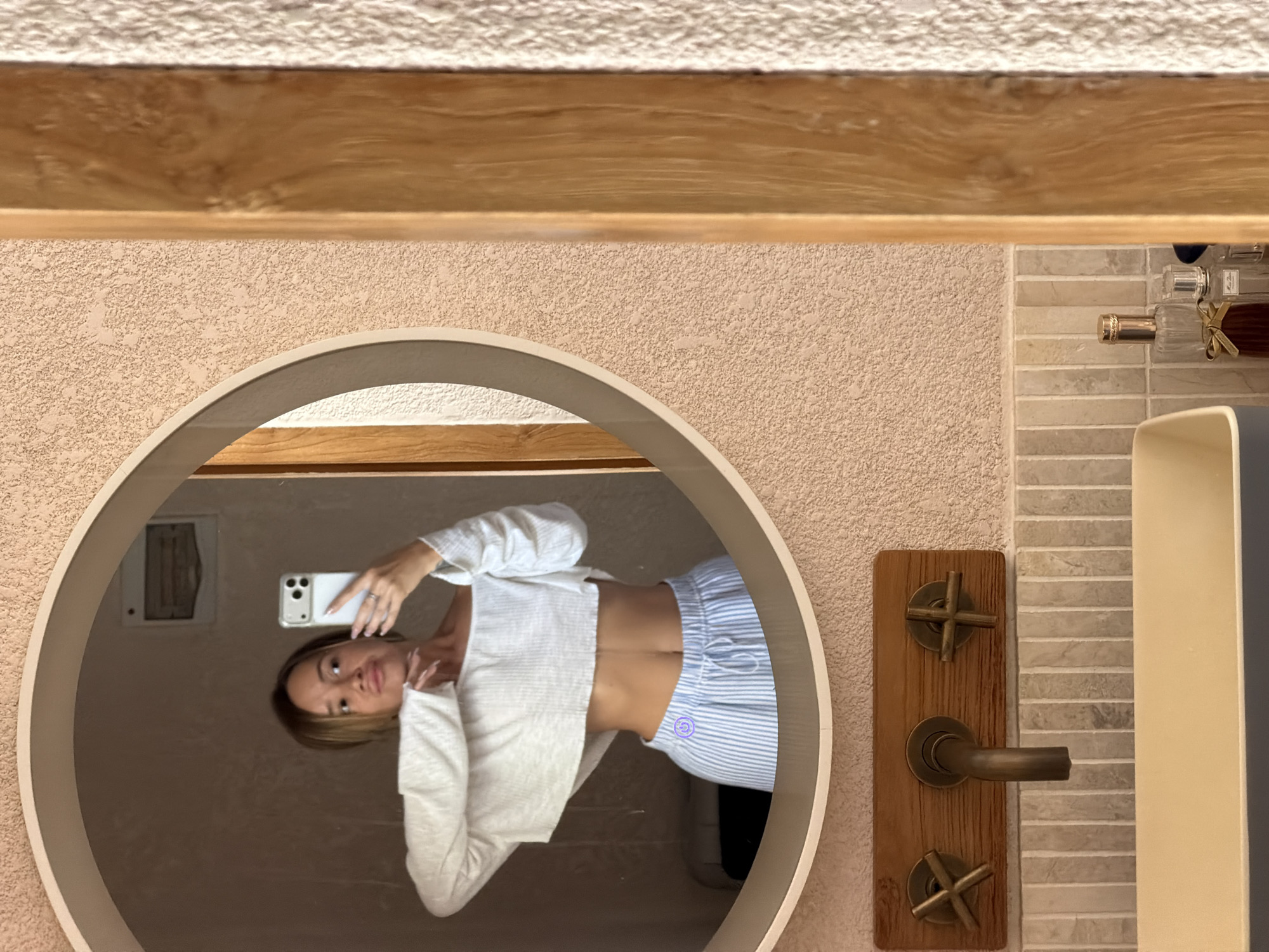 A woman is taking a selfie in a bathroom mirror. The bathroom features textured peach-colored walls, a wooden shelf, and light gray brick tiling with wooden water control knobs and a light gray basin. The woman is reflected in a round mirror, holding a white smartphone up in her right hand. She is wearing a white cardigan and blue and white striped pants. The location of this scene is Mengwi, Indonesia.