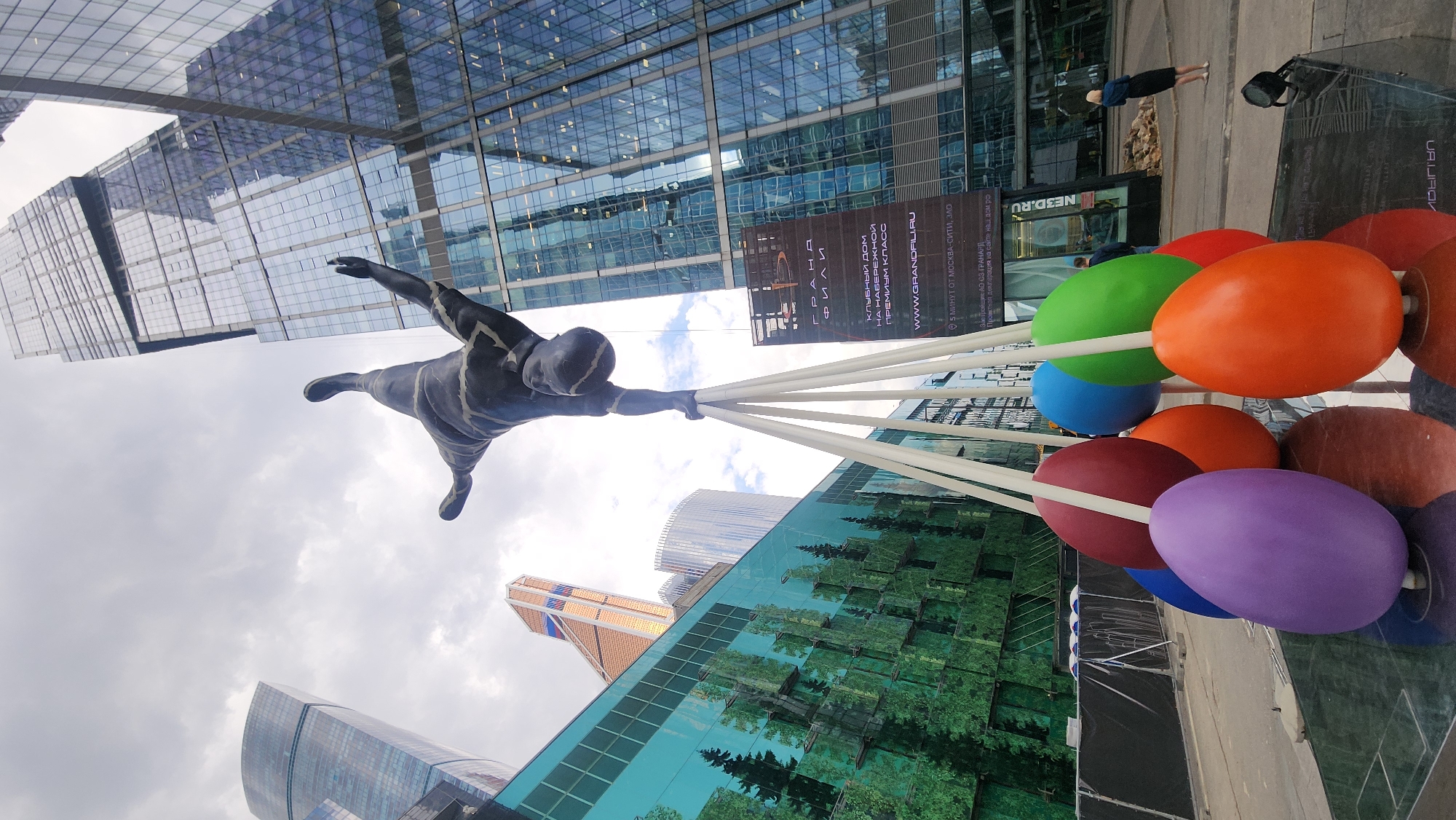 This image showcases a statue of a man holding onto a bunch of colorful, egg-shaped balloons. The location is in the city of Moscow, Russia, near the Moscow International Business Center (Moscow-City), as evidenced by the skyscrapers visible in the background and the inscription mentioning "5 минут от Москва-Сити, ЗЛО".
The statue is positioned so it appears to be flying between the modern buildings. The balloons are multicolored, including shades of red, orange, green, blue, purple, and brown. The camera angle is tilted, giving a dynamic and slightly disorienting perspective of the scene.
A few people can be seen in the background of the image. A sign, part of which is readable as "GRAND FILI" is visible.