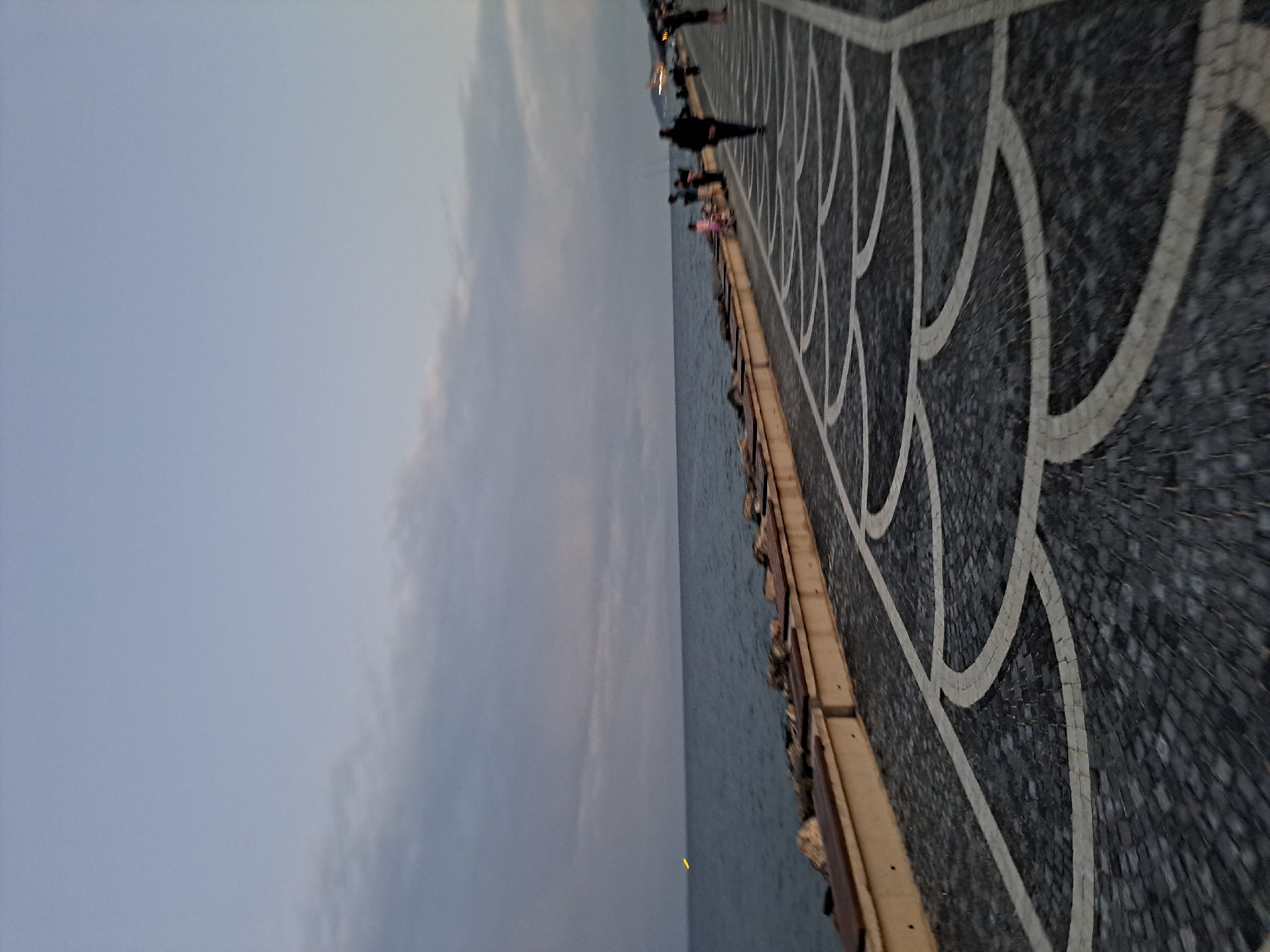 This image captures a scenic view of the Tekirdağ coastline at dusk. The sky is a soft, dusky blue with scattered clouds, hinting at the transition from day to night. The sea is a calm, deep blue, meeting a rocky shoreline with a paved promenade. Along the promenade, several people are visible. Some are walking, while others appear to be standing or engaged in conversation. The promenade itself is paved with dark cobblestones featuring a distinctive white, curved pattern, adding a decorative element to the walkway. In the distance, along the coastline, there are hints of lights, suggesting the presence of buildings or further urban development. A small, yellow light is visible on the water, possibly indicating a buoy or a distant vessel. The overall atmosphere is peaceful and serene, characteristic of an evening stroll along the waterfront.