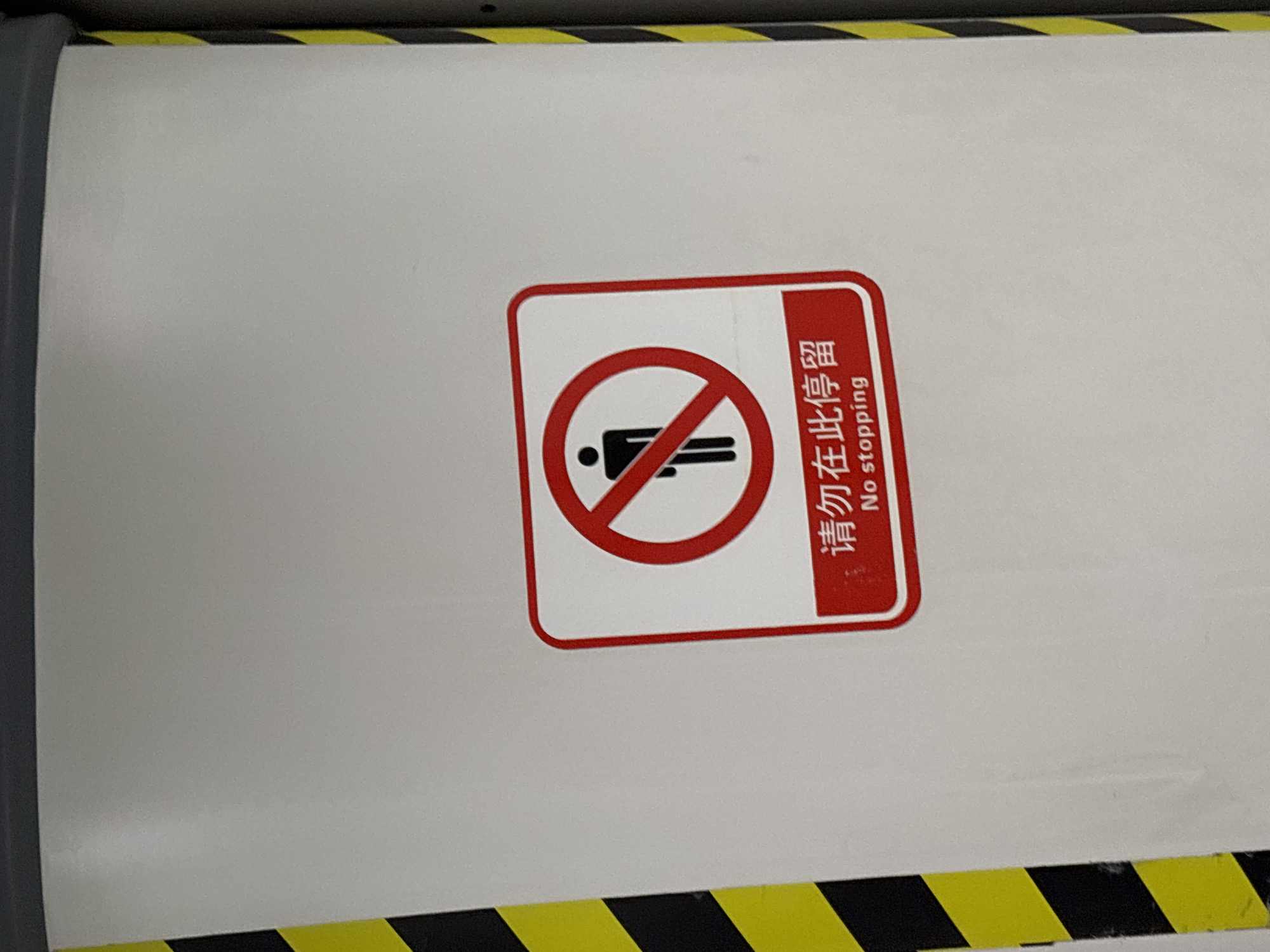The image shows a rectangular white sign with a red border and rounded corners, affixed to a white surface with yellow and black hazard stripes at the top and bottom. The sign displays a red circle with a diagonal line through it, superimposed over a stick figure lying down, indicating "No Stopping". To the right of the symbol, the Chinese characters "请勿在此停留" appear above the English translation "No stopping", both printed in red. The location of the image is potentially in Jiulongpo District, China.