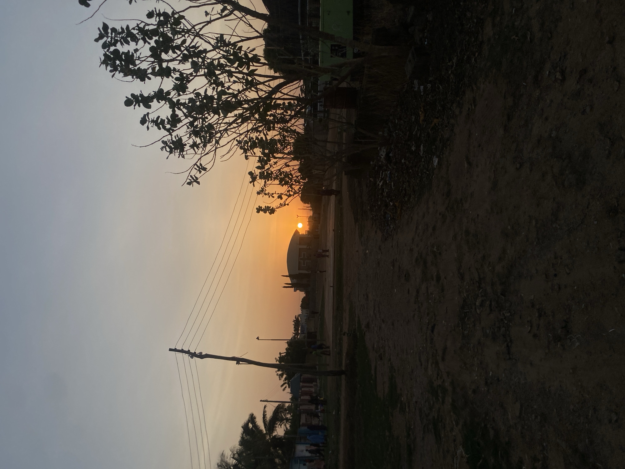 The image depicts a sunset in Barden Yana, Nigeria. The sun is setting over a large building with a rounded roof. A row of trees and utility poles line the road in front of the building. The ground is dirt. There are indistinct figures of people visible in the distance. The foreground is dark.