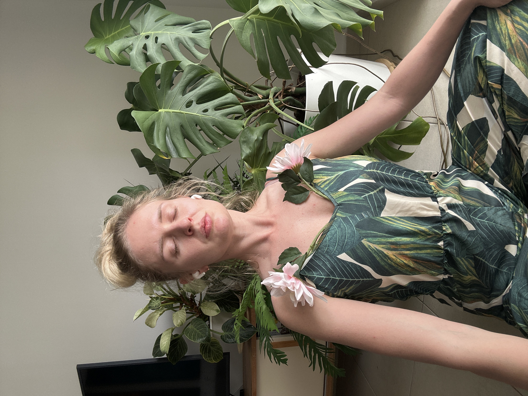 A blonde woman is lying down amid lush green plants and flowers. She wears a jumpsuit with a tropical leaf pattern. There are two pink flowers draped on her chest and shoulder. She has light skin, blonde hair pulled back in a bun, and is wearing wireless earbuds. Her eyes are closed. The plants surrounding her include a large Monstera deliciosa in a white pot. The background includes a grey wall, a black television, and a wooden cabinet. The location cannot be determined from the image and there is no indication this image was taken in Petah Tikva, Israel.