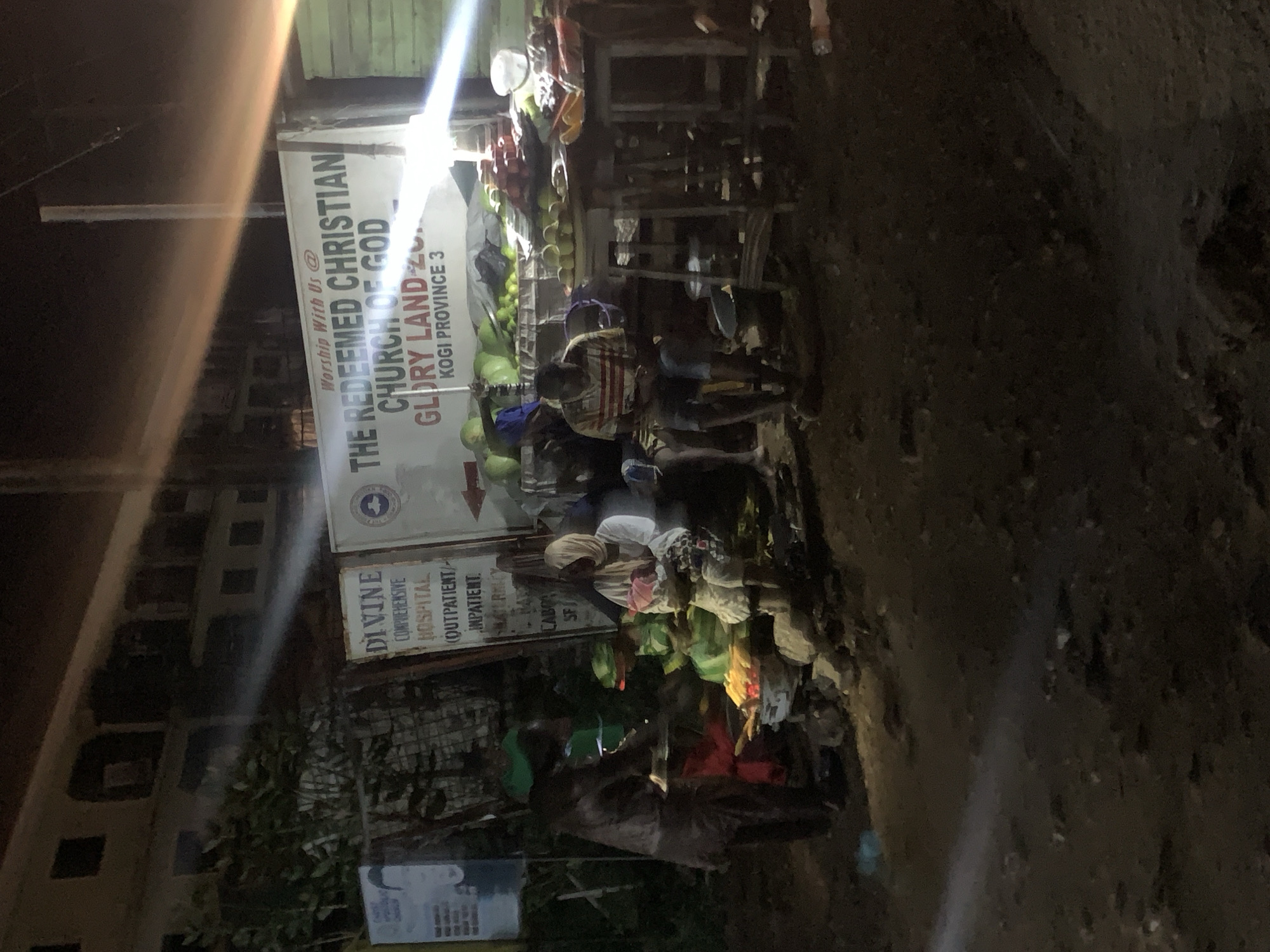 The image depicts a nighttime scene in Lokoja, Nigeria, likely on a street corner where vendors are selling goods. A large banner in the background reads "Worship With Us @ THE REDEEMED CHRISTIAN CHURCH OF GOD GLORY LAND ZONA KOGI PROVINCE 3", indicating the location's affiliation. In the foreground, a group of people are gathered around a makeshift stall where various fruits and produce are displayed. A person in a brown garment is attending to customers, possibly serving food or taking orders. Another individual, wearing a striped t-shirt and shorts, is seated and looking down, while a woman in a hijab sits nearby with a child. The ground appears to be dirt and uneven, with scattered debris. The lighting is dim, with artificial lights from the stall and possibly streetlights illuminating the scene, creating strong contrasts and shadows. There are also signs in the background, one of which mentions "DIVINE COMPREHENSIVE HOSPITAL". The overall impression is of a bustling, informal marketplace operating in the evening.