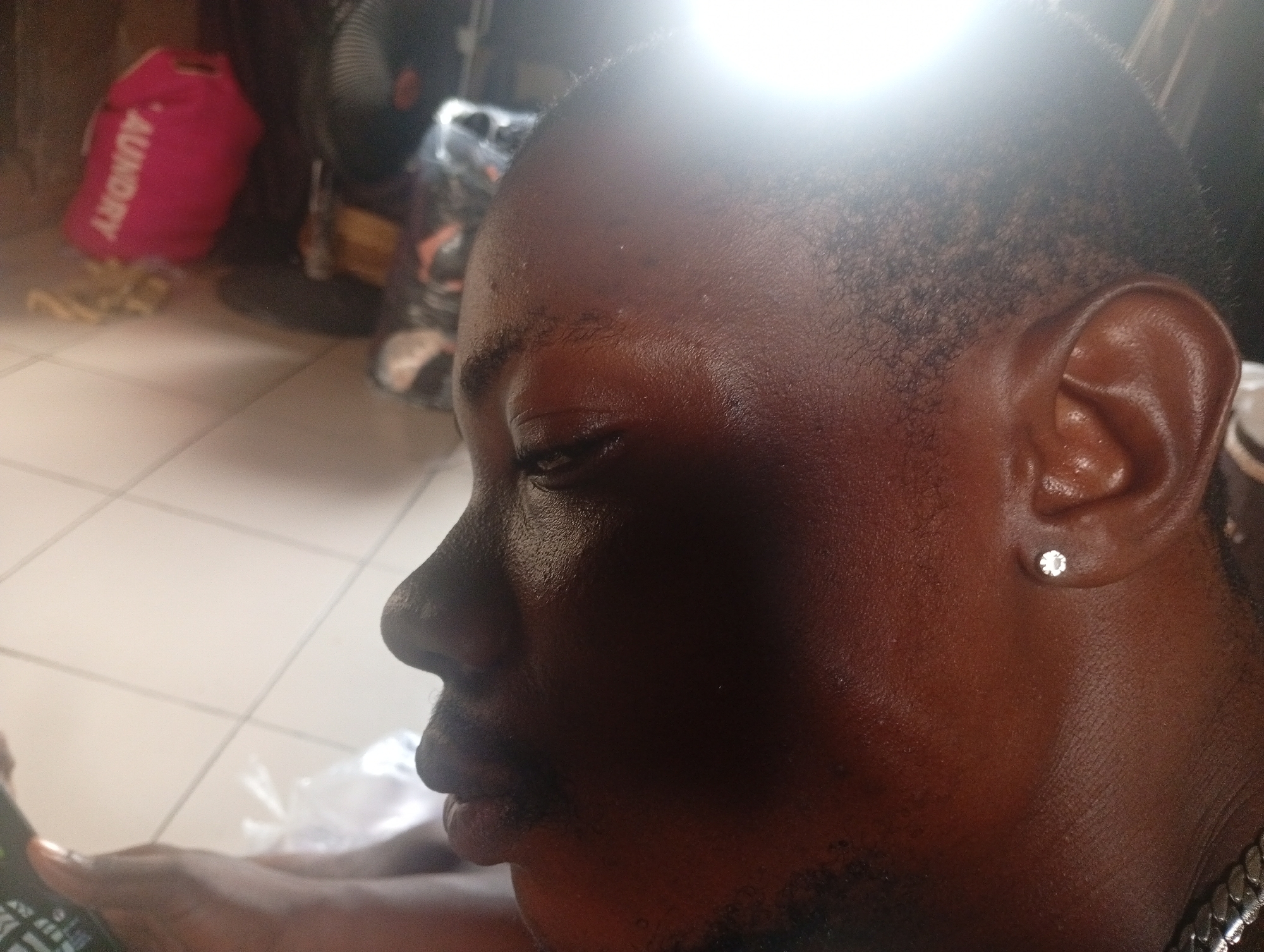 The image shows a close-up, side profile of a Black man's face. He is wearing a stud earring in his right ear. The lighting is bright, creating a lens flare at the top of the image. In the background, a pink laundry bag with "LAUNDRY" written on it in white letters is visible, along with other indistinct objects and a fan. The man appears to be indoors, possibly in a home or a room with a tiled floor. The scene is not in a city, but rather an indoor setting.