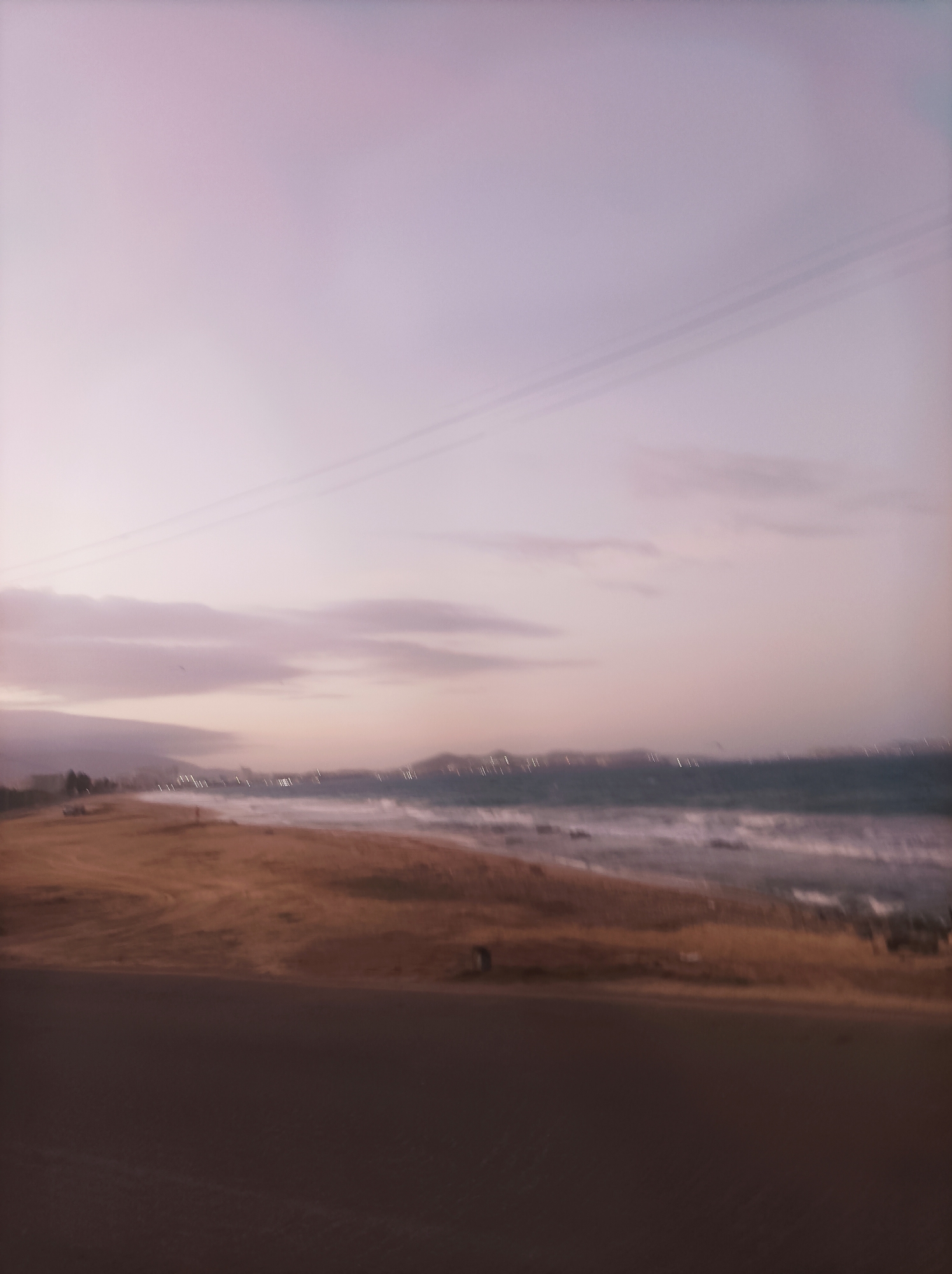 The image shows a blurry view of a beach in Porlamar, Venezuela, at twilight. The sky is a soft gradient of pink and purple hues with faint clouds. There's a dark road or paved area in the foreground, and a sandy beach extends toward the blurred coastline. The ocean waves are visible, breaking along the shore. In the distance, there are blurred lights from the city along the coast. Above, two power lines run across the sky.