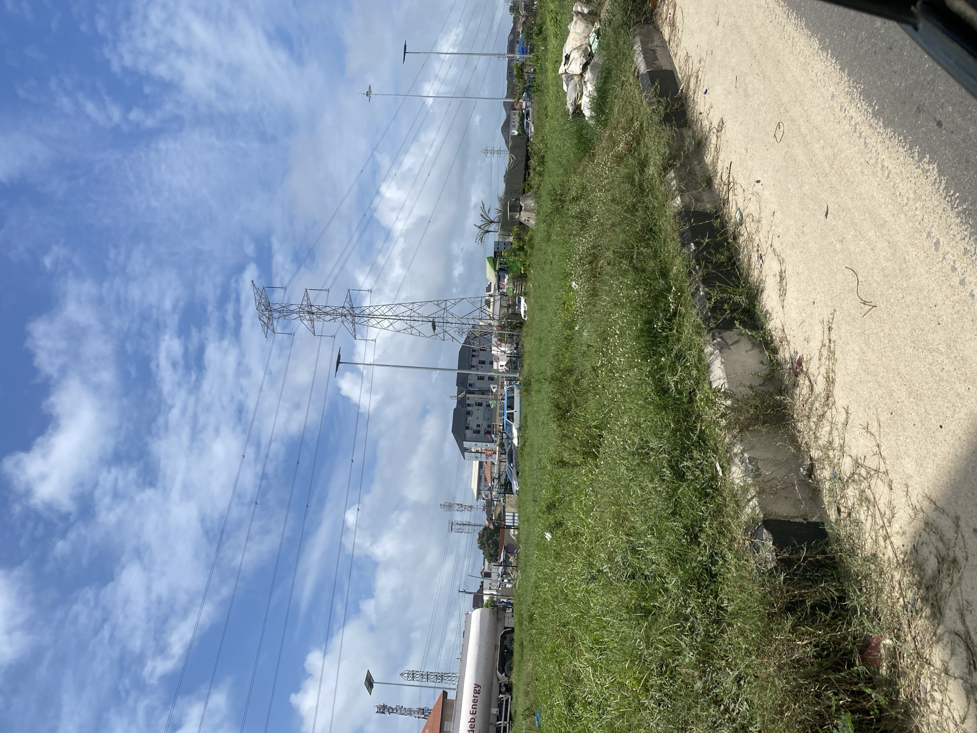 The image captures a scene on the Lekki Peninsula II in Nigeria, featuring a wide, paved road in the foreground with an unkempt grassy verge. The verge is lined with a concrete curb that is partially overgrown with weeds and debris. Beyond the verge, several vehicles are visible on a road, including a white and blue van, cars, and a large white tanker truck labeled "Jdeb Energy." In the background, numerous tall electrical pylons and overhead power lines dominate the skyline, interspersed with various buildings of different sizes and architectural styles, some appearing to be residential or commercial. The sky is bright blue with scattered white clouds. Several tall, slender light poles are also present along the roadside. The overall impression is one of an active urban environment with significant infrastructure.