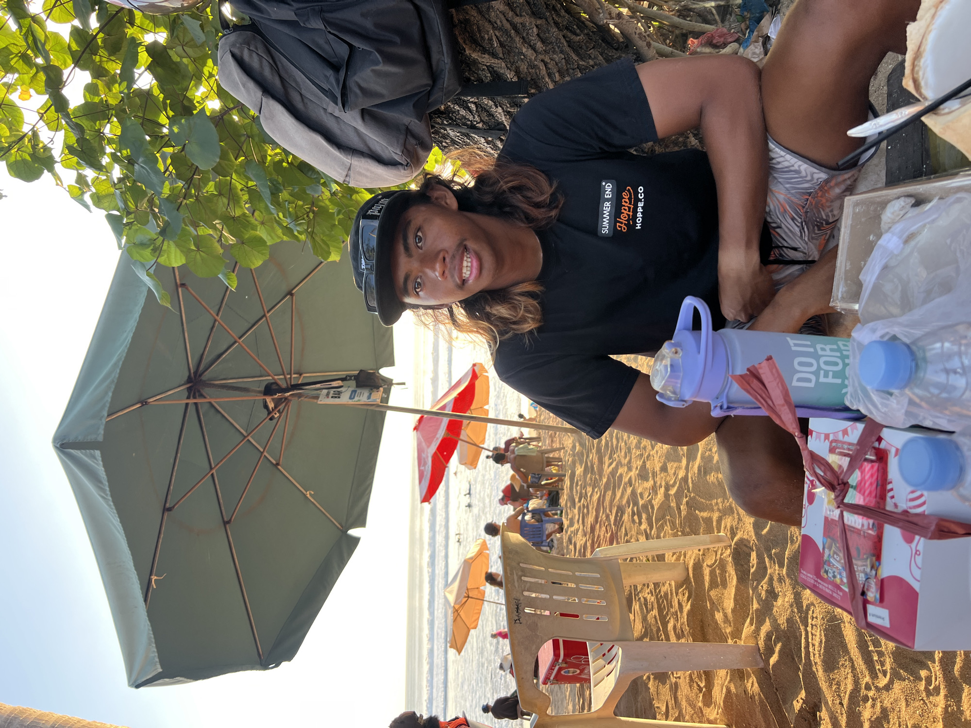 A medium shot captures a young man sitting on a sandy beach in Kuta, Indonesia, as the sun sets. He wears a black t-shirt with an orange logo that reads "SUMMER END Hoppe HOPPE.CO", and a black baseball cap. The man has long, wavy brown hair and is looking towards the left of the frame.
In the foreground, a table holds several items: a pink and white box with red ribbon, a blue water bottle, and two plastic bottles of water. A small white box is also visible. Behind the table, beach chairs and umbrellas dot the sandy expanse. In the background, gentle waves roll onto the shore, and several people are visible in the ocean, some surfing. The overall lighting suggests a warm, late afternoon or early evening. The environment is a typical beach scene with sand, umbrellas, and the ocean.