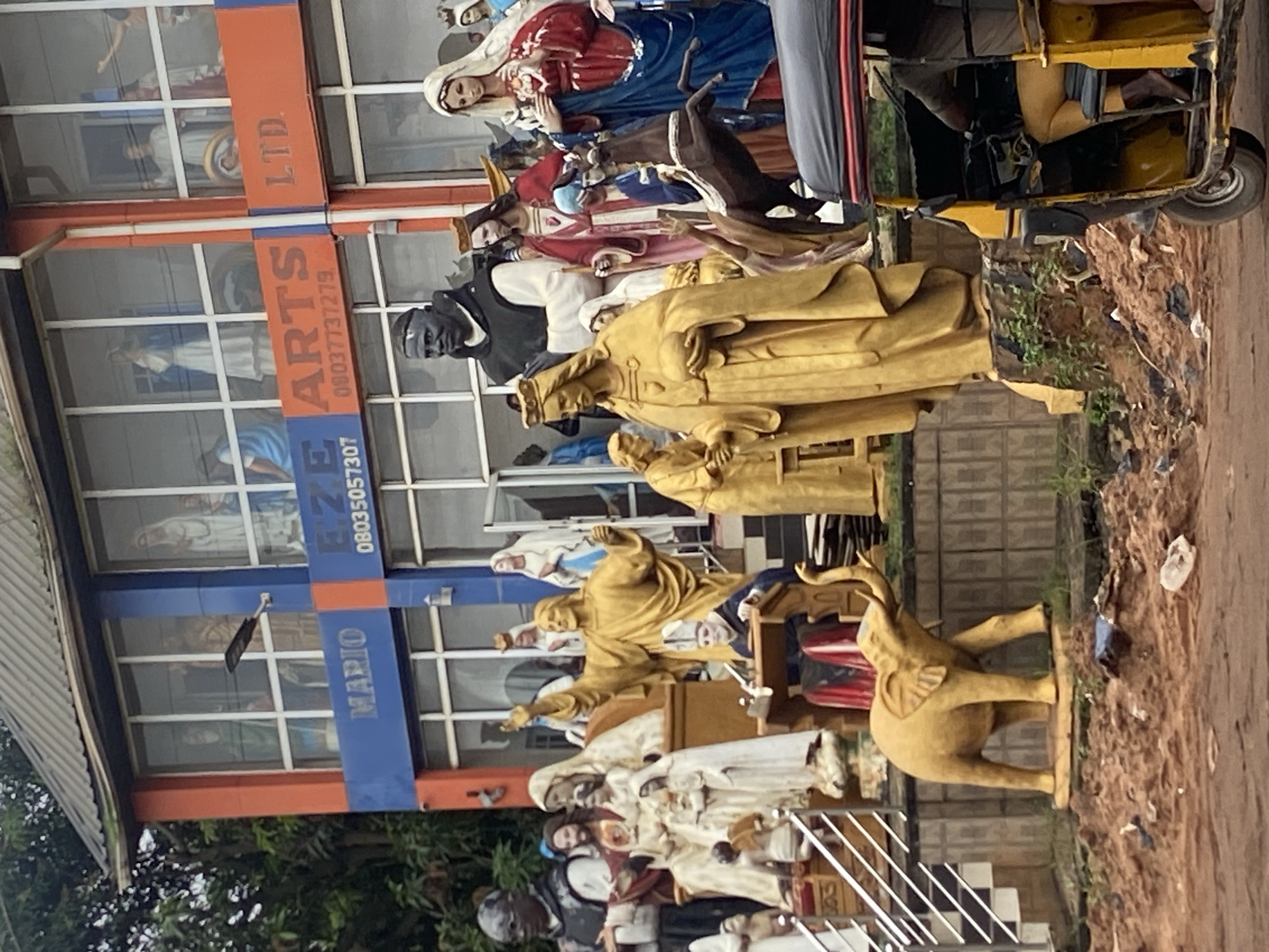 In Nkpor, Nigeria, a vibrant scene unfolds in front of a multi-story building adorned with blue and orange signage. The building displays the names "MARIO" and "EZE ARTS LTD." and a phone number "08035057307." The facade features numerous windows, each framing statues of religious figures, predominantly in white and gold.
In the foreground, a collection of religious statues commands attention. A golden statue of Jesus, arms outstretched, stands prominently. To its right, another golden statue depicts a king or religious leader holding a sphere. To the left of the Jesus statue, a figure in a dark robe and white habit, resembling a monk, is visible. Several other white statues of women in traditional attire are also present.
Further to the left, a large, golden elephant statue stands. Behind the statues, a yellow auto-rickshaw, a common mode of transport in Nigeria, is parked. A person, partially visible, is seated in the driver's seat. The ground in front of the building is uneven and covered with reddish-brown earth, debris, and sparse vegetation. The overall impression is a lively commercial area with a strong religious theme.