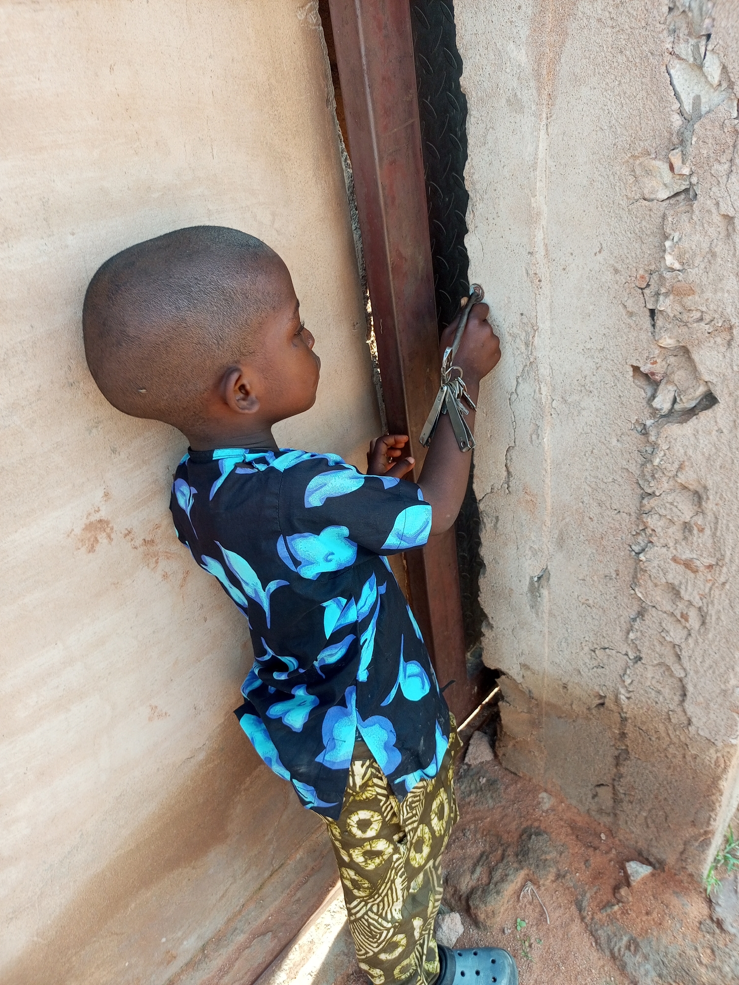 A young child with a shaved head, wearing a blue and black floral print shirt and patterned pants, stands with their back to the camera, reaching towards a rough, light-colored wall with a set of keys in their right hand. The child is positioned next to a dark metal structure, possibly a door frame or gate. The environment appears to be outdoors, with a dirt and gravel ground. The wall shows signs of age and damage, with cracks and exposed materials. The image is taken from a high angle, looking down at the child. The setting is likely a residential area in Tempure, Nigeria.