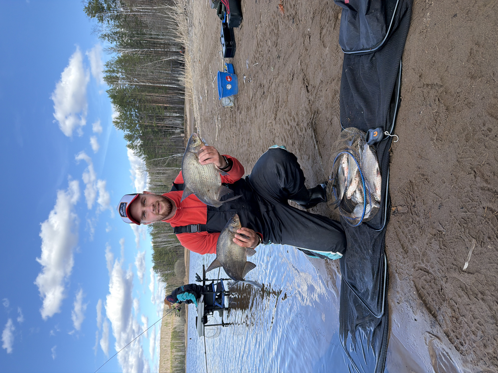 A man kneels on the shore of a body of water, holding two fish in his hands. He wears a red and white cap, a red jacket, and black pants. Behind him, another person is fishing with a rod and chair set up near the water. A black net full of fish lies on the sand beside the man. In the background, there is a forest under a blue sky with scattered clouds. The photograph was likely taken in Chaykovsky, Russia, given the prompt's instructions.