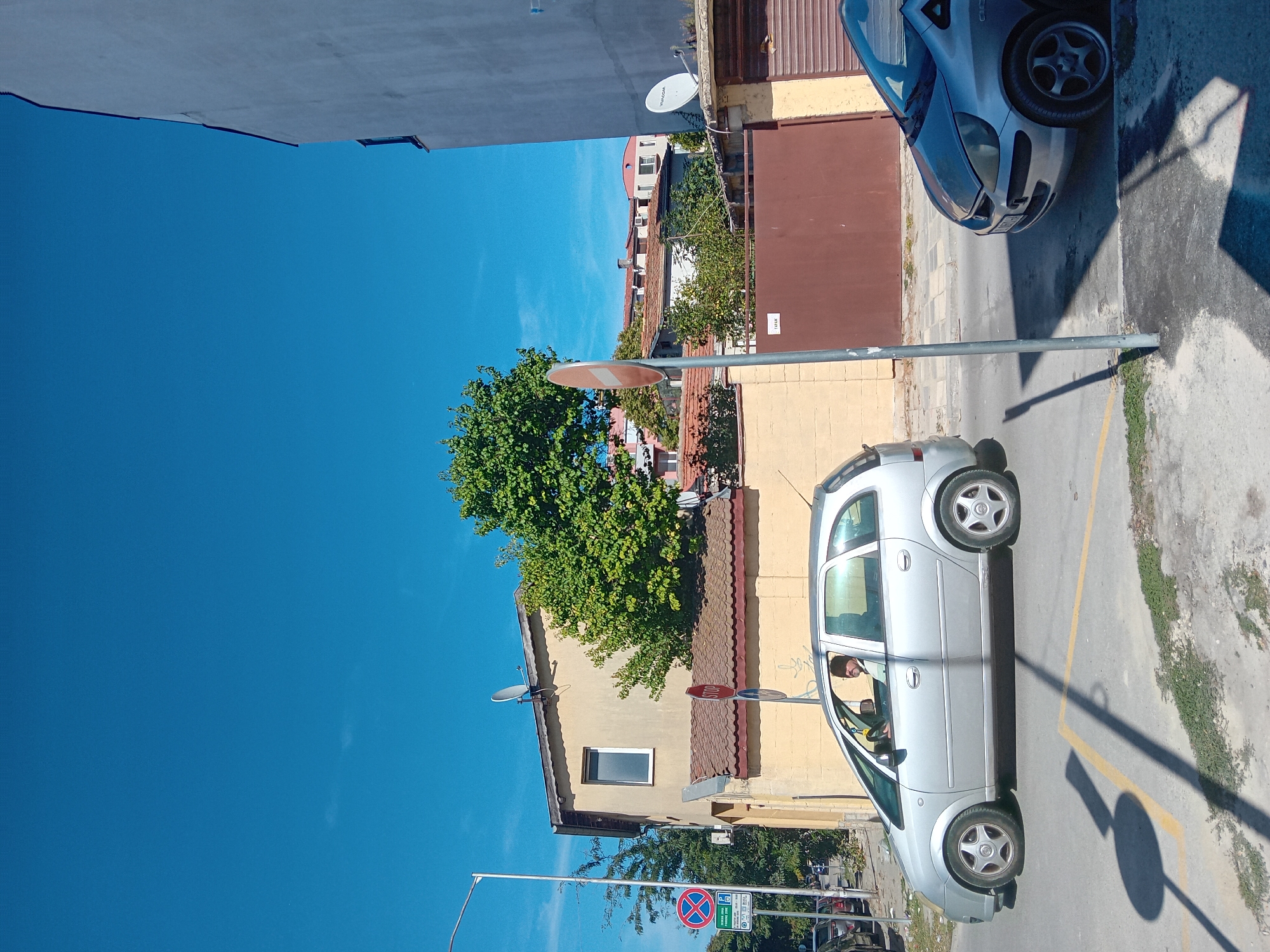 This is a ground-level, eye-level, outdoor, street-level, medium shot of a street scene in Varna, Bulgaria. The day is bright and clear, with a vibrant blue sky. In the foreground, on the right side of the image, a silver compact car is parked, facing left. A silver minivan is in motion on the left side of the street, moving away from the camera. A man with dark hair and a beard, wearing a light-colored shirt, is visible driving the minivan. Yellow lines mark the road, indicating a lane or parking area. A grey utility pole with shadows cast across the street stands in the middle ground. Behind the minivan, a light yellow building with a brown tiled roof and a small white-framed window is visible. A large green tree with dense foliage is to the right of this building. Further in the background, several other buildings with red tiled roofs and some satellite dishes are scattered across the horizon. Traffic signs are present, including a circular red and white "STOP" sign and a blue circular sign with a red diagonal line, indicating no parking. A darker brown gate with a sign reading "ТАРАН" is also visible. The overall scene depicts a typical urban street in Varna on a sunny day.