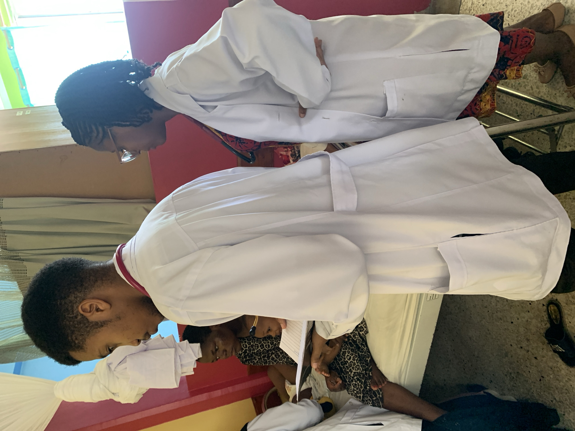 Two individuals in white lab coats are examining a patient. One individual is wearing glasses and has braided hair. The other individual has short hair and a red object around their neck. The patient is sitting on a hospital bed, and a woman is seated next to the patient. The location appears to be a medical facility in Dar es-Salaam, Tanzania.