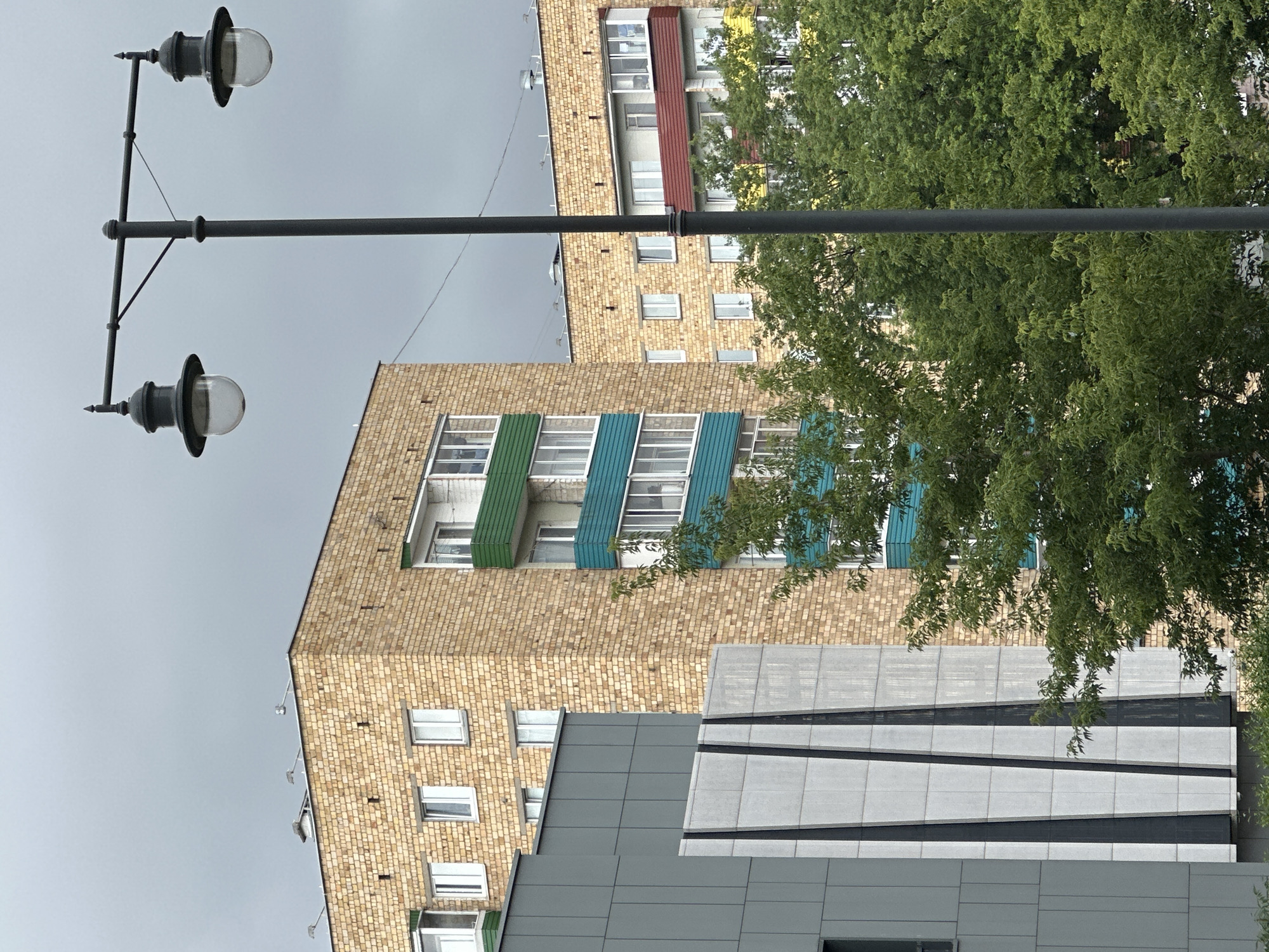 The image depicts a street scene in Minsk, Belarus, under an overcast sky. In the foreground, a dark grey lamppost with two ornate, globe-shaped lights stands prominently. Behind the lamppost, a modern building with a grey facade and geometric patterns is visible on the left. To the right of this structure, a multi-story brick building with beige brickwork and colorful balconies (green, teal, red, and yellow accents) rises. Trees with lush green foliage partially obscure the lower sections of the buildings. The architecture suggests a residential area, typical of urban development in Eastern European cities. No people are present in the frame.