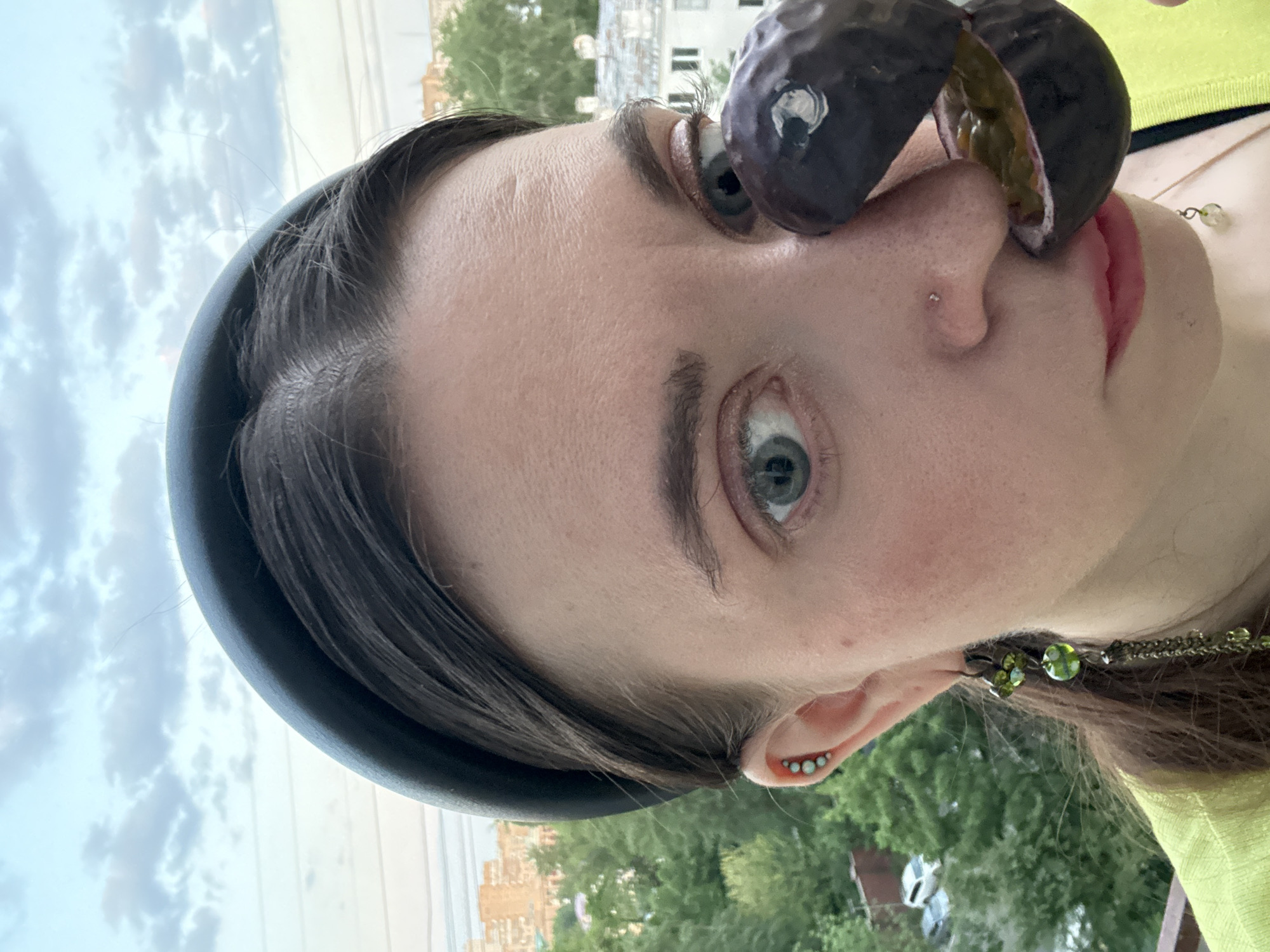 A young woman with fair skin, wearing a black headband and a neon yellow top, holds a halved passion fruit against her nose in an outdoor setting. She has light blue eyes and a nose piercing. The background shows trees, buildings, and a cloudy sky, indicating an urban area. Based on the architecture visible, it's potentially Moscow, Russia.