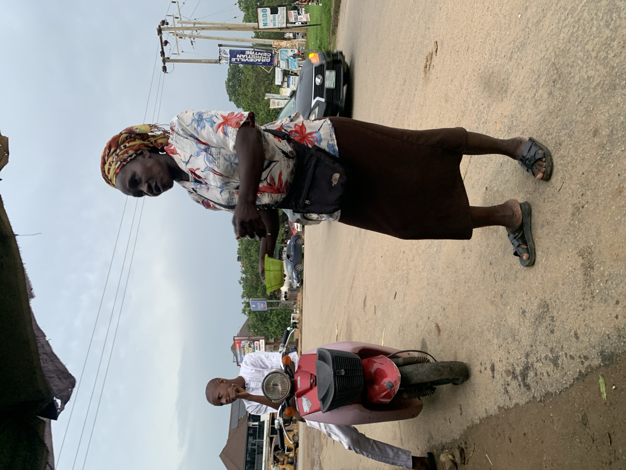 In Abuja, Nigeria, a street scene unfolds with two individuals and a red and pink scooter. A woman, wearing a floral-patterned shirt, a brown skirt, and a headscarf, stands on the right side of the frame, holding a green bowl in her hands. A young boy, dressed in a white traditional outfit, sits on the scooter, looking to his left. The scooter is parked on the side of a paved road, with other vehicles and buildings visible in the background. Power lines and street signs are also present, indicating an urban environment. The sky is overcast, suggesting a cloudy day.