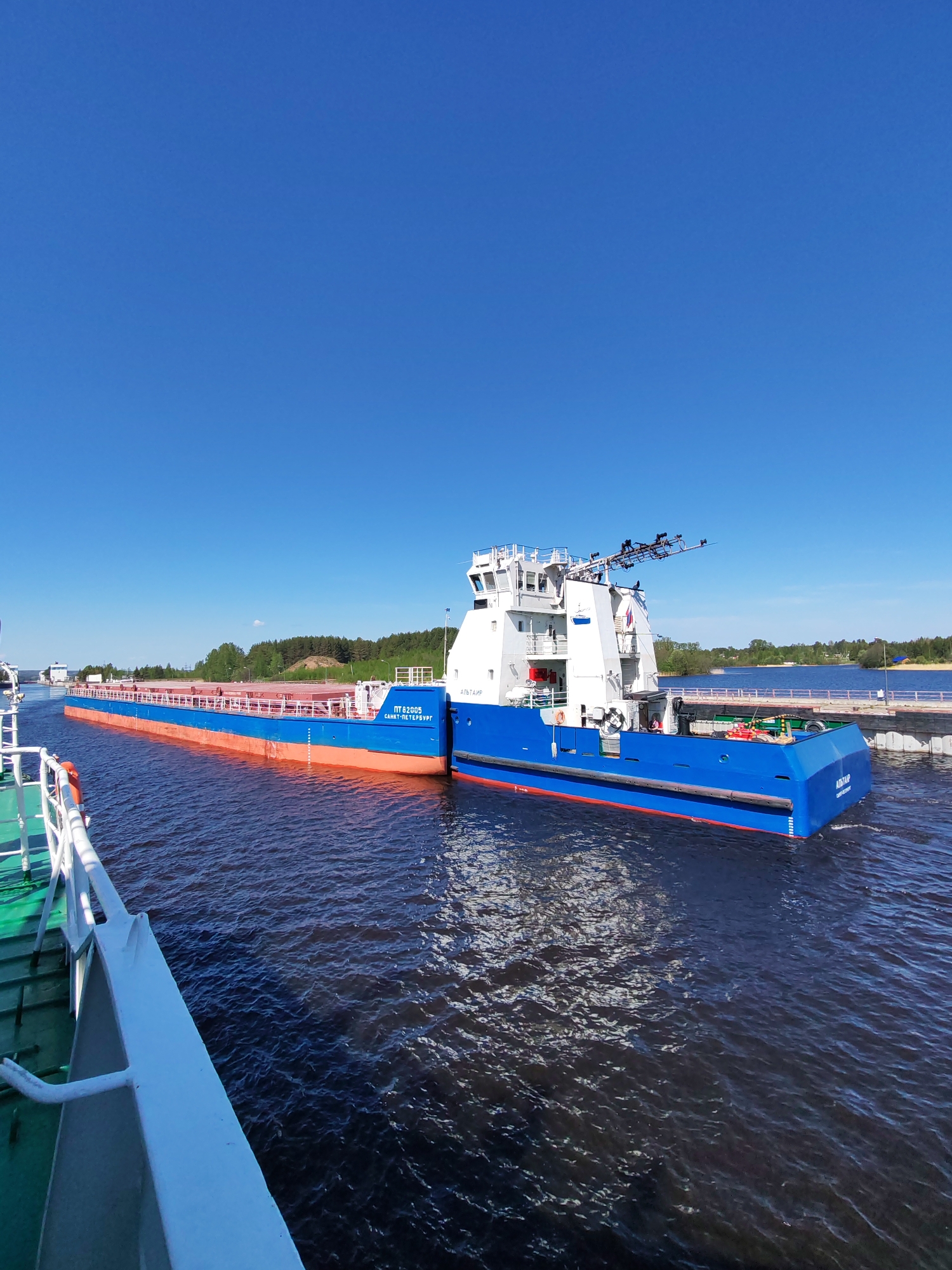 The image captures a scene on a waterway, possibly a canal or river, in Белоусово, Russia. The main subject is a blue and white barge-like vessel named "АЛЬТАИР," with visible Russian text "ПТ 628005 САНКТ-ПЕТЕРБУРГ" on the side, indicating its registration in Saint Petersburg. The barge has a flat cargo area. The front portion of another boat is visible on the left side of the image, with green and white paint. The water is dark and choppy. In the background, there is a bridge with a visible light and a wooded shoreline. The sky is clear blue, indicating bright daylight conditions.