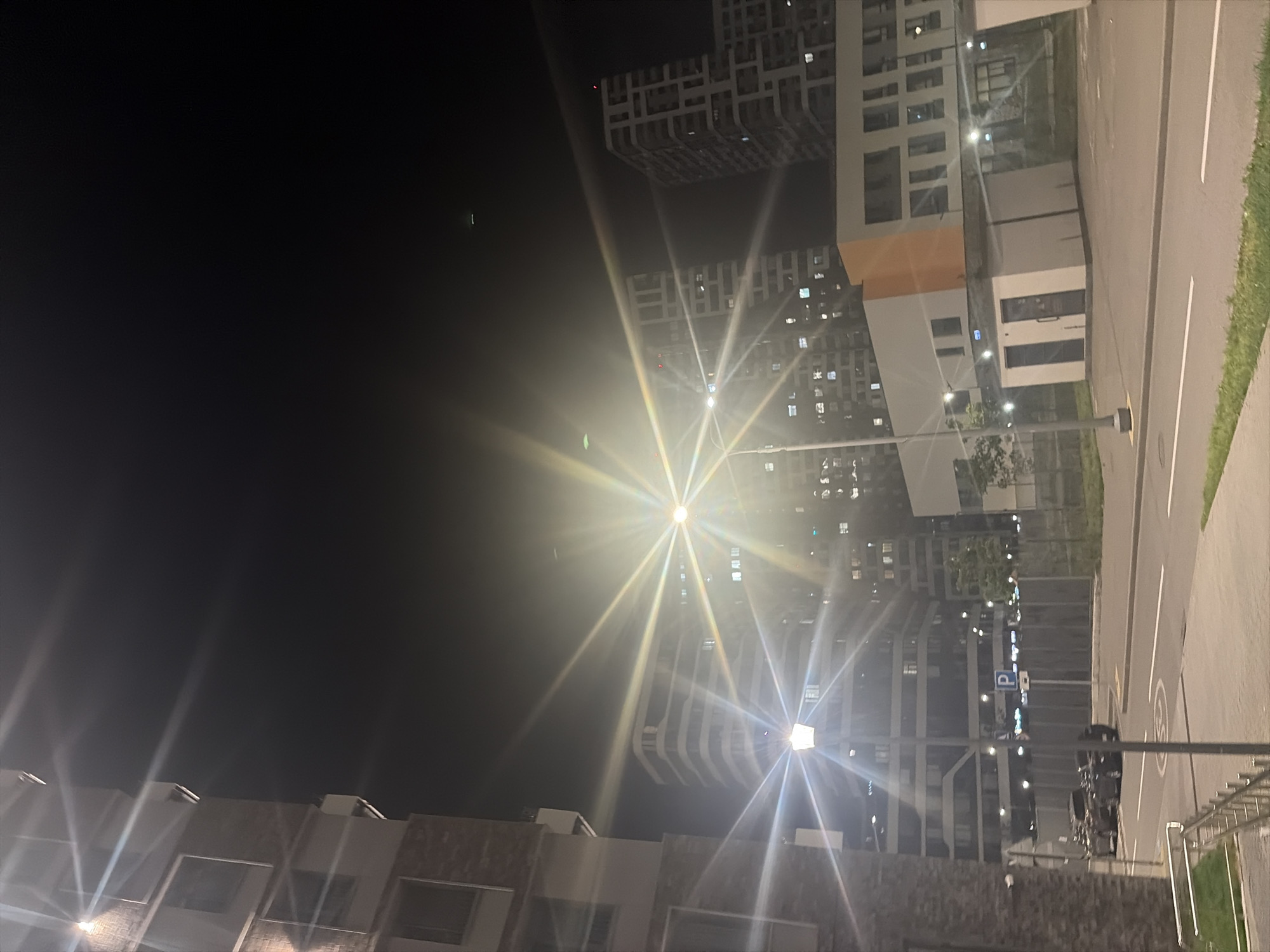 An exterior, low-angle, medium shot captures a nighttime scene in a modern urban environment, likely in Moscow, Russia, characterized by tall residential buildings. A prominent streetlamp in the foreground emits a bright, starburst effect due to lens flare, illuminating the surrounding area. To the left, the textured facade of a brick building is visible. In the mid-ground, several high-rise residential buildings with numerous illuminated windows create a backdrop. A low-rise building with orange and white accents is also present. In the foreground, a paved road runs horizontally, with parked cars and a blue parking sign visible on the right side of the street. The sky is dark and clear.