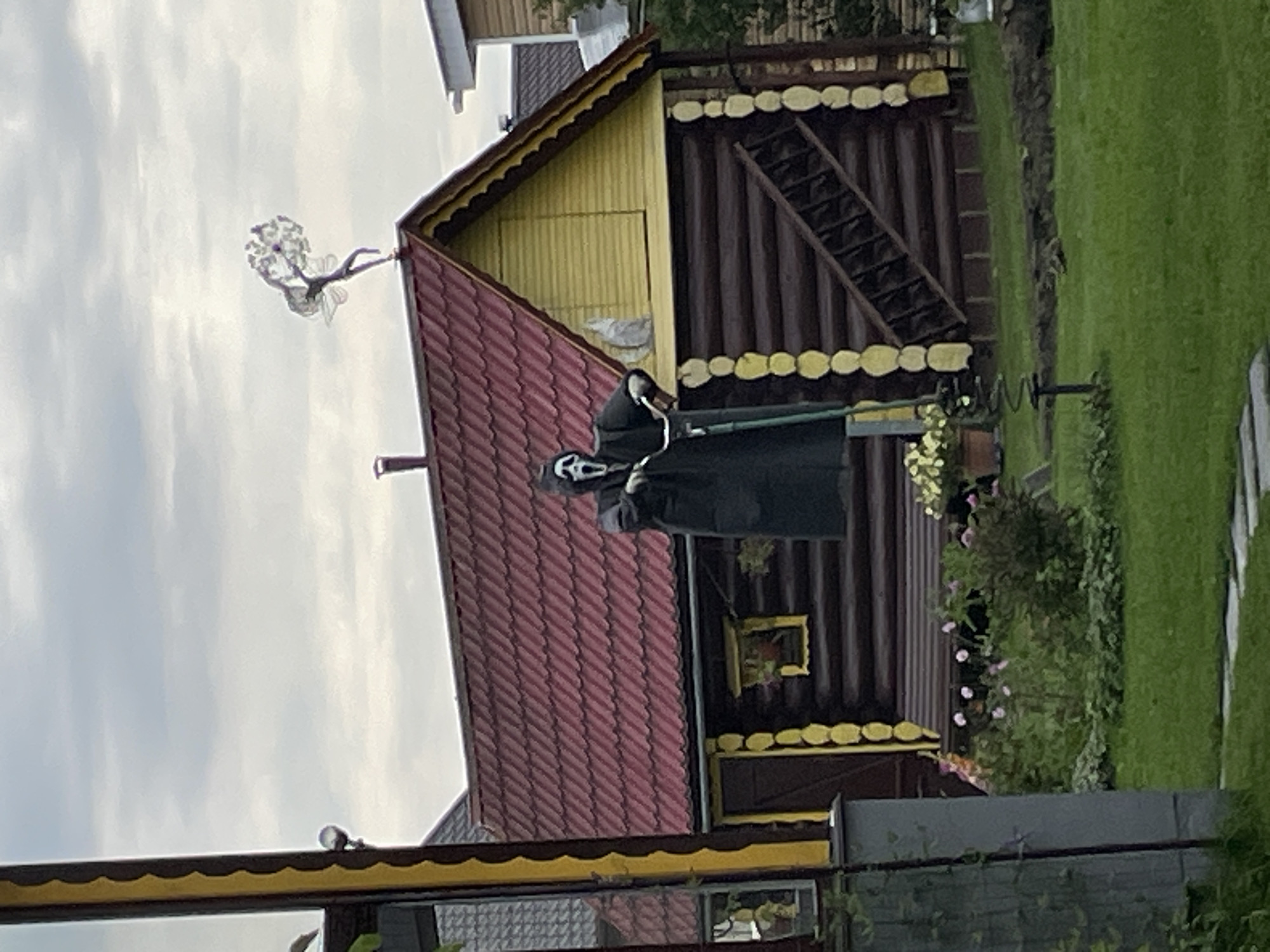 The image depicts an outdoor scene with a log cabin-style building under a cloudy sky. In front of the building, a figure dressed in a black cloak and a white Ghostface mask from the Scream movie franchise is posed on a scooter. The figure is positioned on a raised platform or porch area in front of the cabin.
The cabin itself has a red, shingled roof and yellow trim around the gabled section. The walls are constructed from dark logs, with lighter colored decorative elements between them. A small window with a yellow frame is visible on the cabin wall. To the left of the cabin, a portion of another building, possibly a house or shed, with yellow and brown vertical siding is visible.
In the foreground, a green lawn covers the ground, with a stone pathway leading towards the cabin. There are several potted plants and bushes with pink flowers scattered in front of the cabin. A thin, dark pole with a spiral spring extending upwards is located on the lawn to the left. On the roof of the cabin, a decorative metal sculpture of a figure with wings is mounted.
The overall impression is one of a residential or garden setting, possibly a dacha (summer house) in a rural or suburban area. The location is identified as Строймаш Снт, Russia. The weather appears overcast.