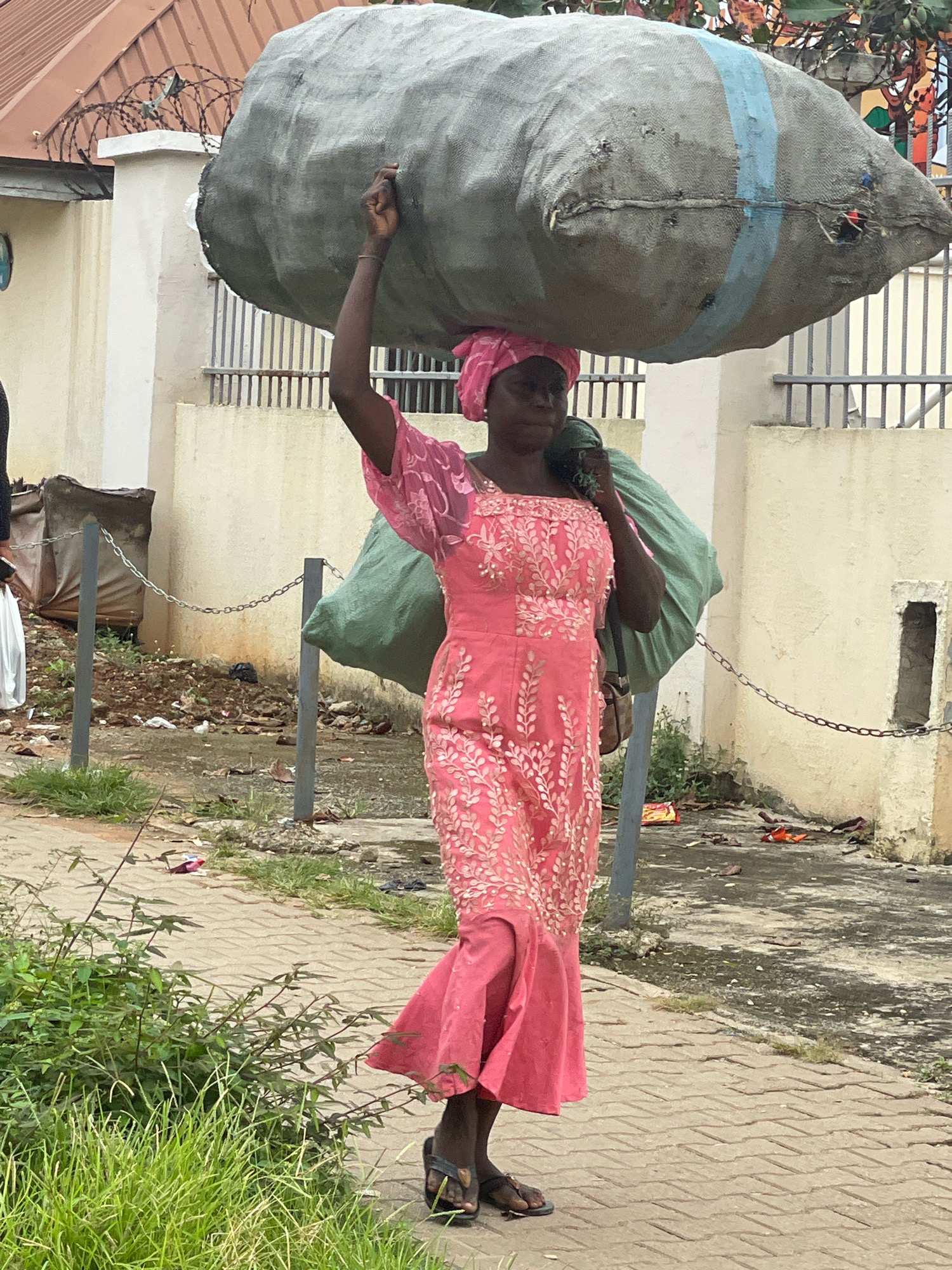 A woman wearing a pink dress and headscarf walks on a paved walkway. She is carrying a large, grey, sack-like object on her head and another green bag over her right shoulder. The woman is walking from right to left in the frame. Behind her, there is a wall with a metal fence in front of it. There are some discarded items and vegetation on the ground near the wall. The scene appears to be an outdoor urban environment, possibly in Abuja, Nigeria, given the context.