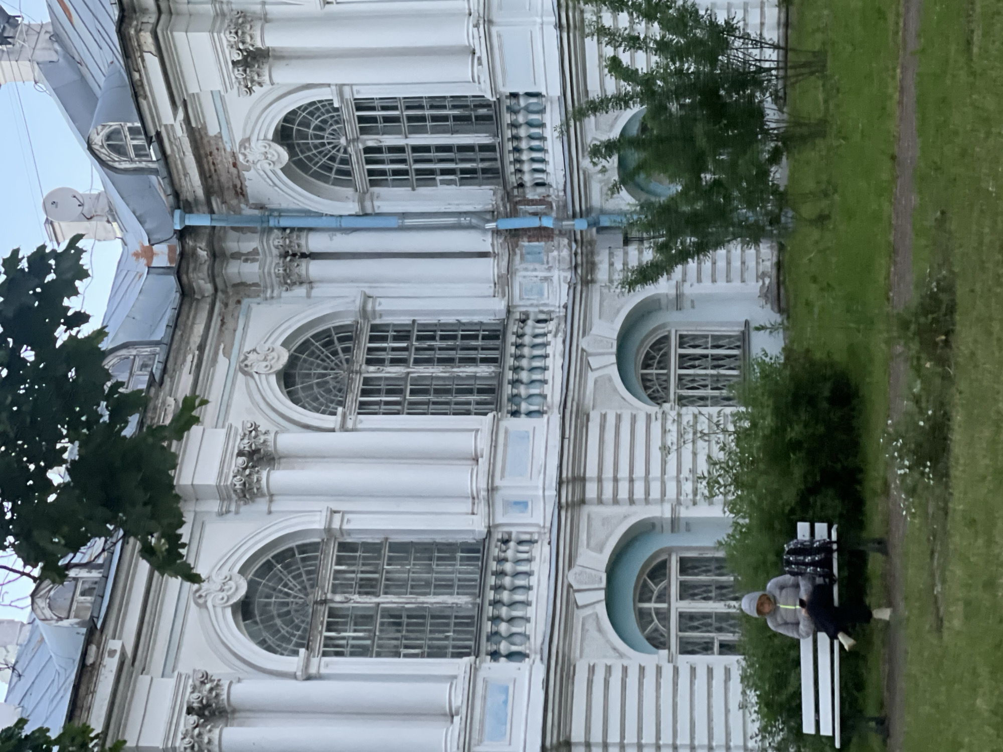 A person sits on a white bench in front of a large, ornate, white building with blue accents. The building has large, arched windows with intricate panes and decorative columns. A blue downspout runs down the side of the building. Green foliage, including trees and bushes, is visible in the foreground and around the building. The person is wearing a grey hooded jacket and dark pants and is looking down at their phone. A patterned backpack is next to them on the bench. The scene appears to be in a park or garden setting. The building's architecture suggests it is likely a historic structure. This is likely located in Saint Petersburg, Russia, given the architectural style.
