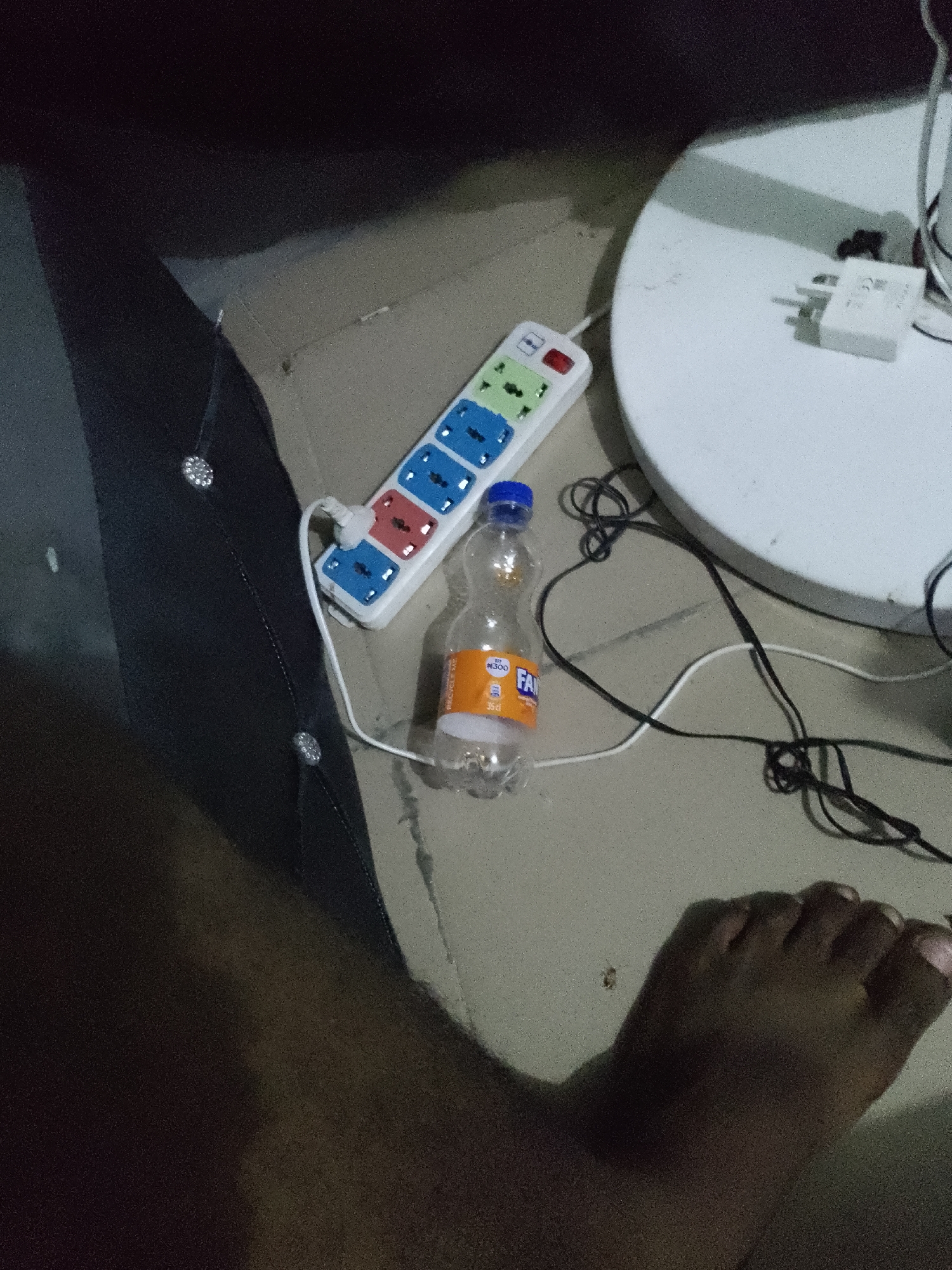 In an indoor environment, a multi-socket power strip is plugged into an electrical outlet. A clear plastic bottle with an orange label, appearing to be a soft drink, rests next to the power strip. Black and white electrical cords are visible, some connected to the power strip and others extending out of frame. A white, round object, possibly a table or stool, is present in the background, with a white electrical adapter plugged into its surface. A person's foot is visible in the foreground, suggesting a human presence in the scene. The floor is tiled or concrete.
